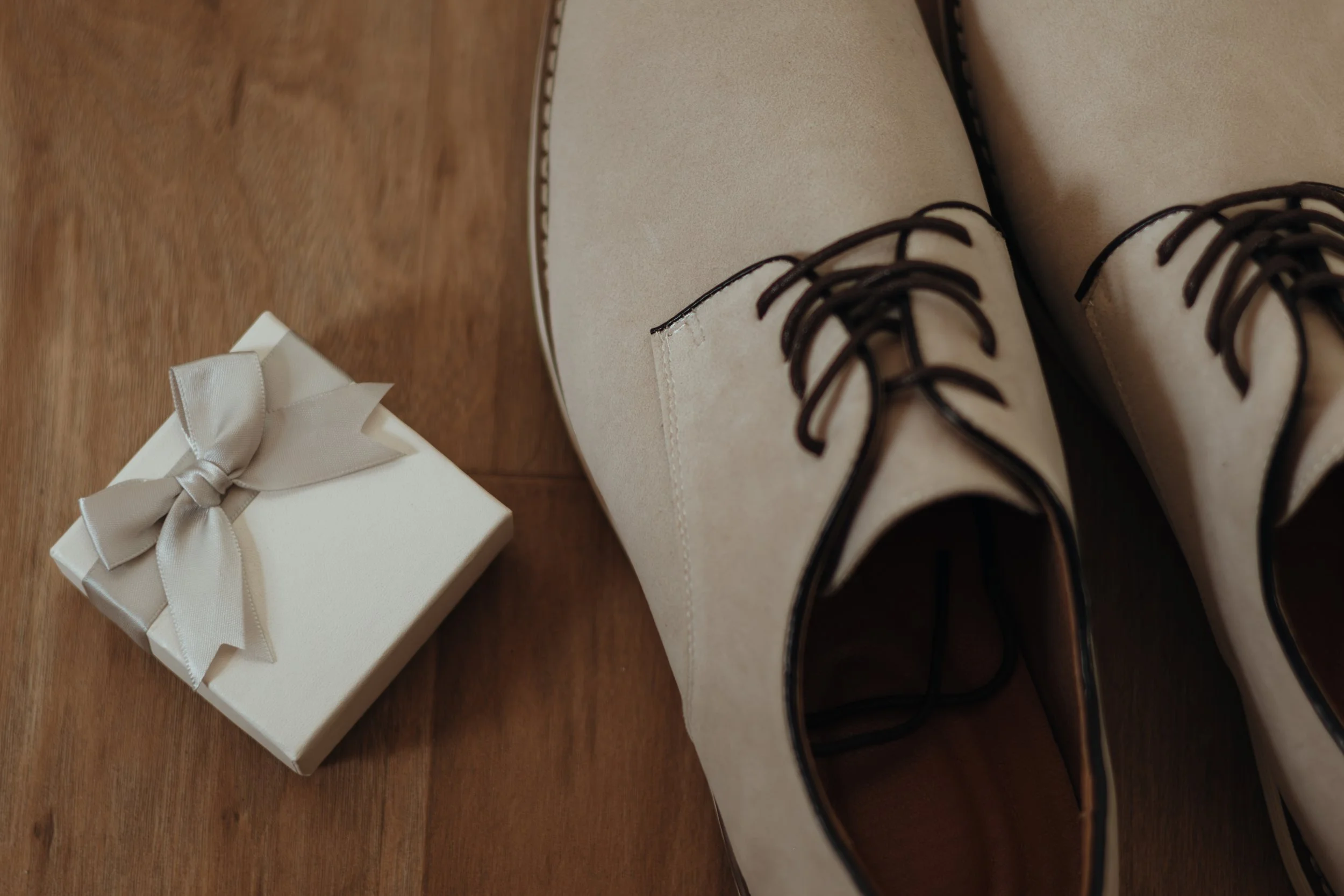 Ein Paar weiße Schuhe mit schwarzen Schnürsenkeln neben einer weißen Geschenkbox mit silberner Schleife auf einem Holzboden.