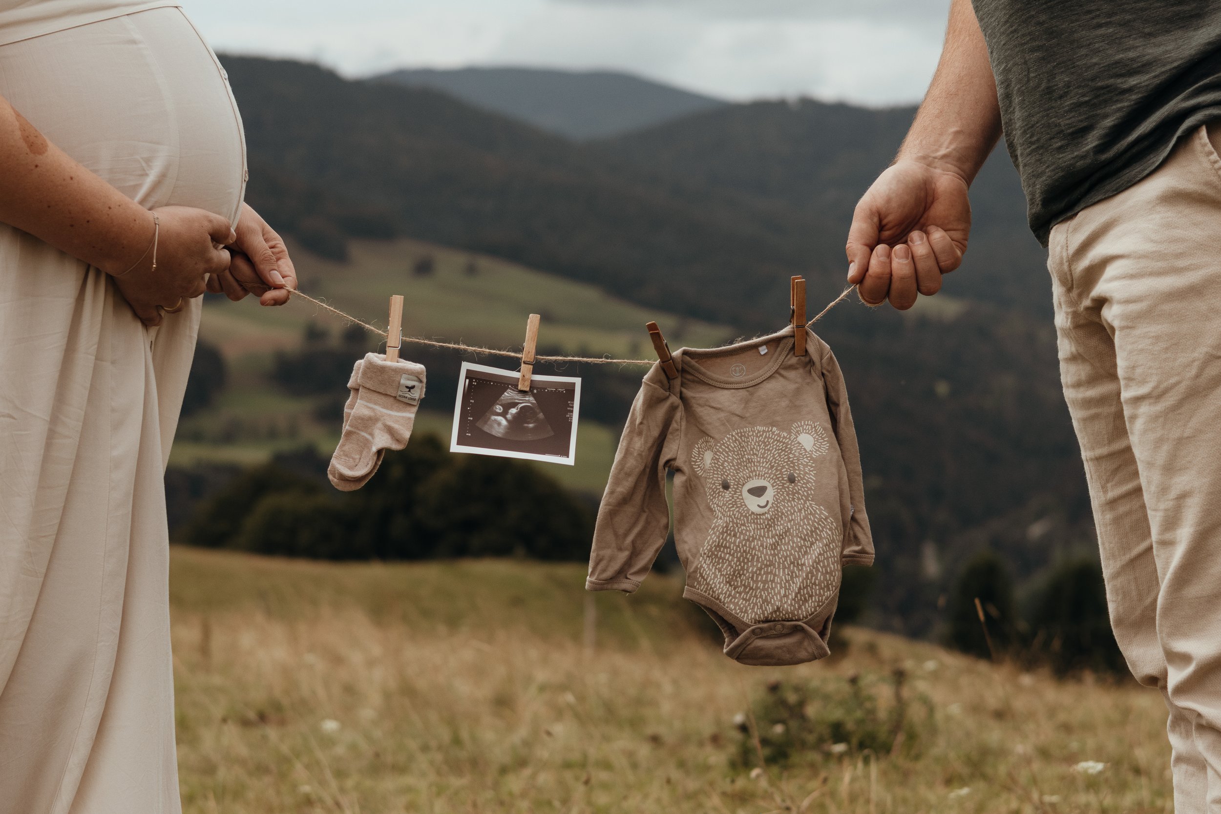 Ein Paar hält eine Wäscheleine mit einem Schwangerschaftsstreifen, einem Ultraschallbild, einem Babystrampler mit Bärenmuster und einem Babystrumpf in einer ländlichen Umgebung.