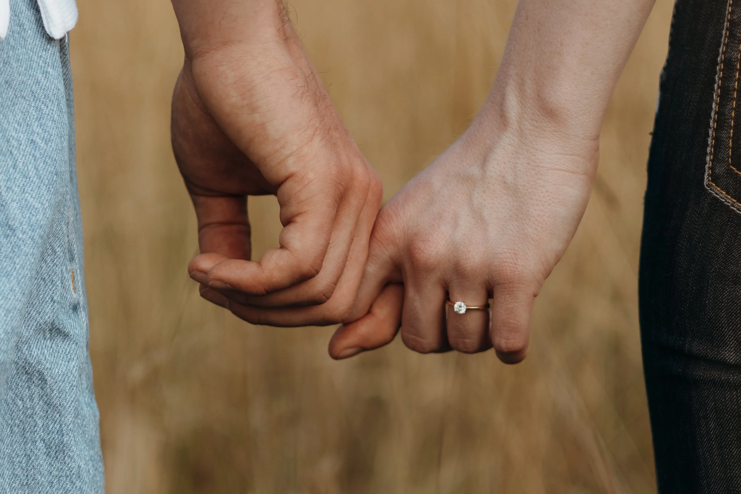 Nahaufnahme von zwei Händen, die sich halten. Eine Hand trägt einen Verlobungsring mit einem Diamanten.