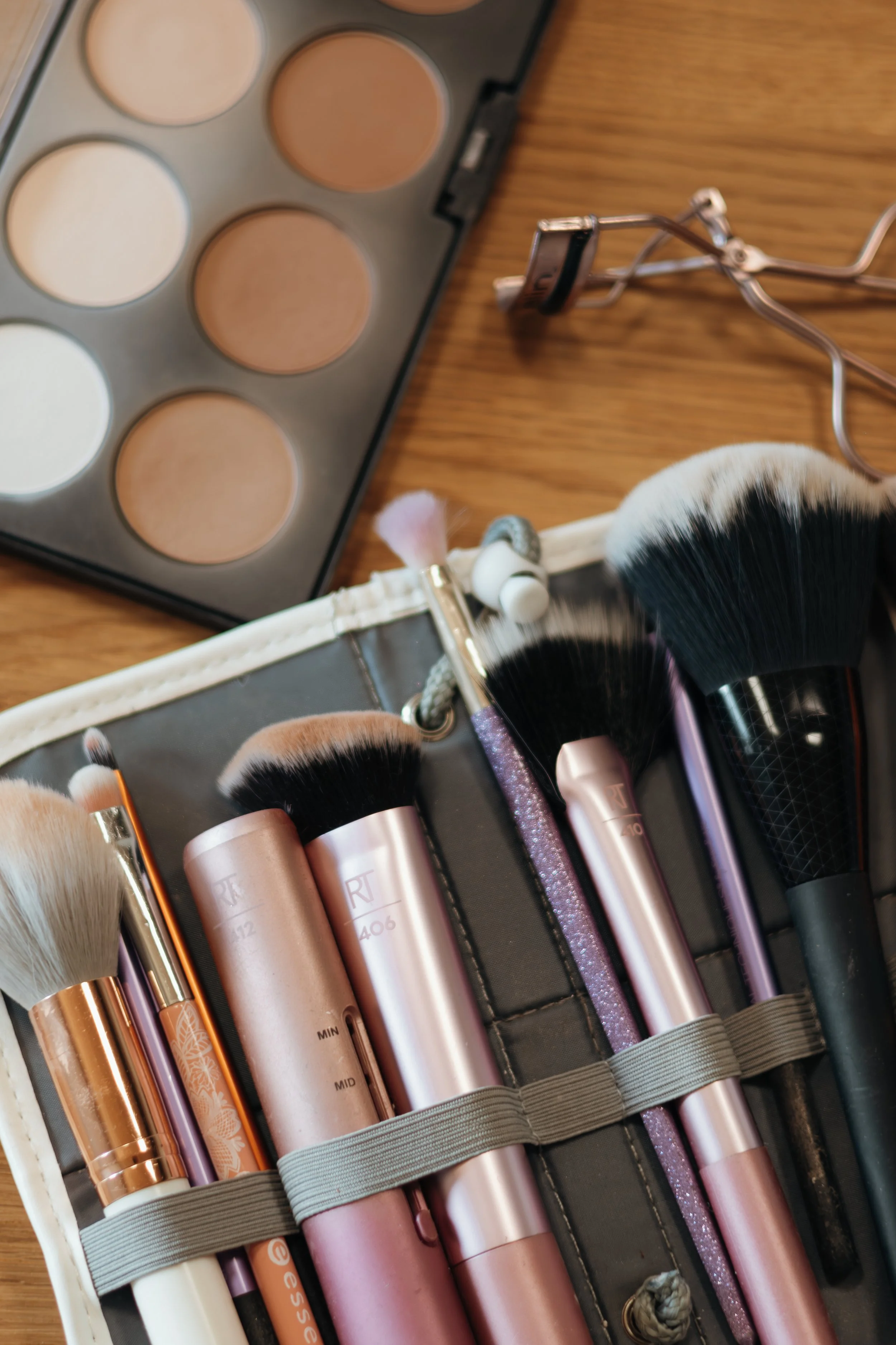 Makeup-Pinsel, Puder, und Wimpernzange auf einem Schminkkasten mit Foundation im Hintergrund.