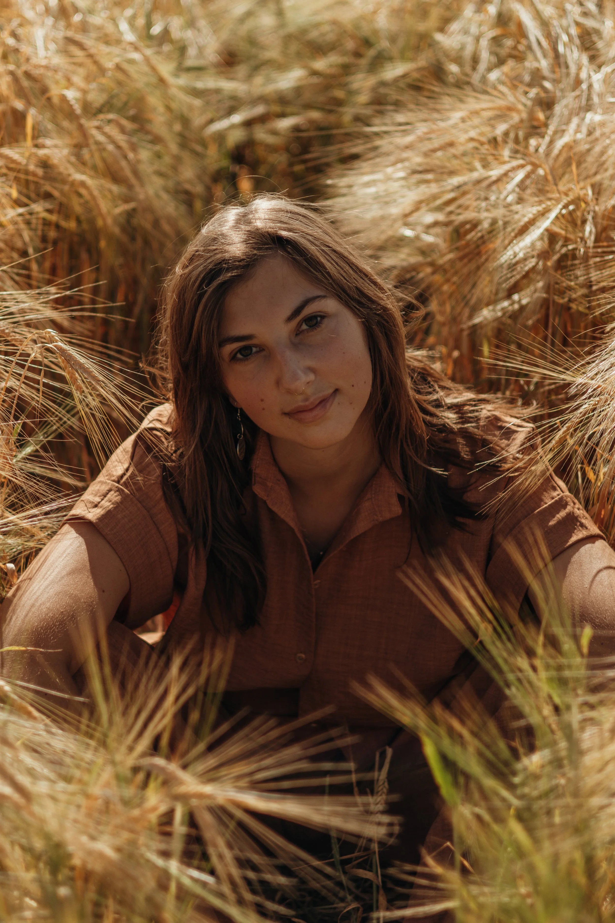 Junge Frau liegt in einem Weizenfeld, blickt in die Kamera, trägt ein braunes Hemd, sonniges Wetter.