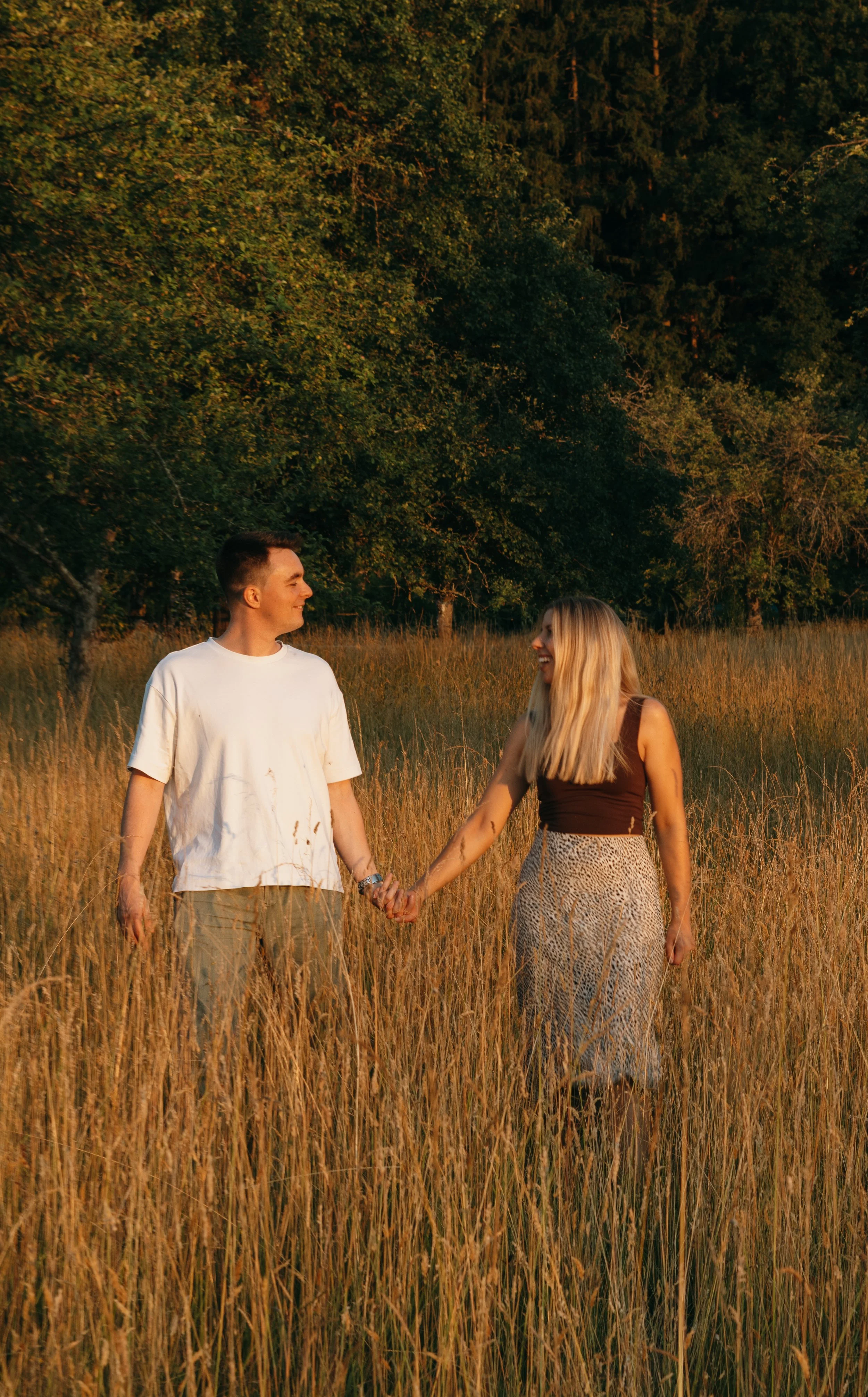 Ein Paar hält Händchen in einem Feld bei Sonnenuntergang, umgeben von Bäumen, sie lachen und schauen sich liebevoll an.