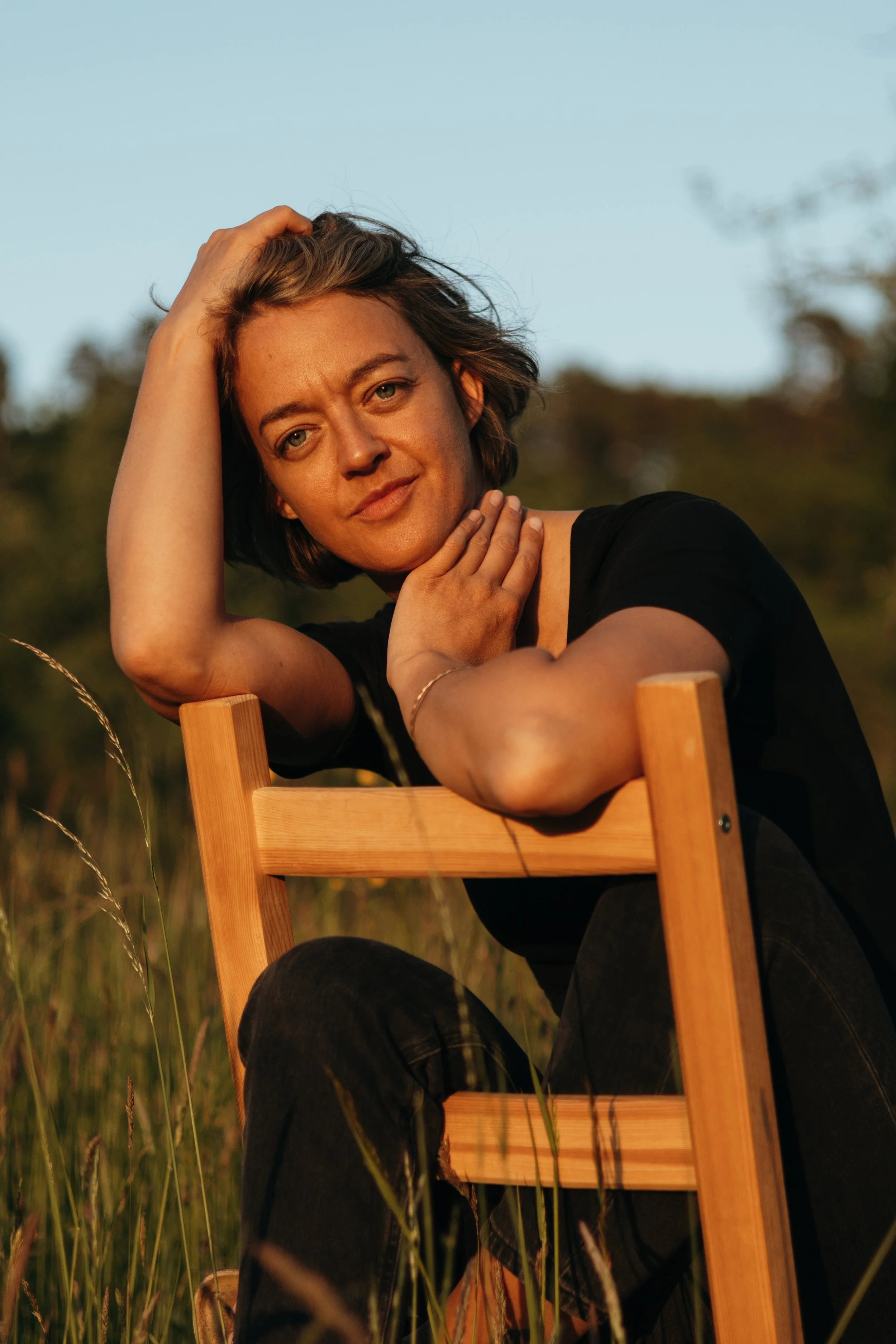 Ein junge Frau mit braunen Haaren sitzt auf einem Holzstuhl im Freien, während es sonnig ist.