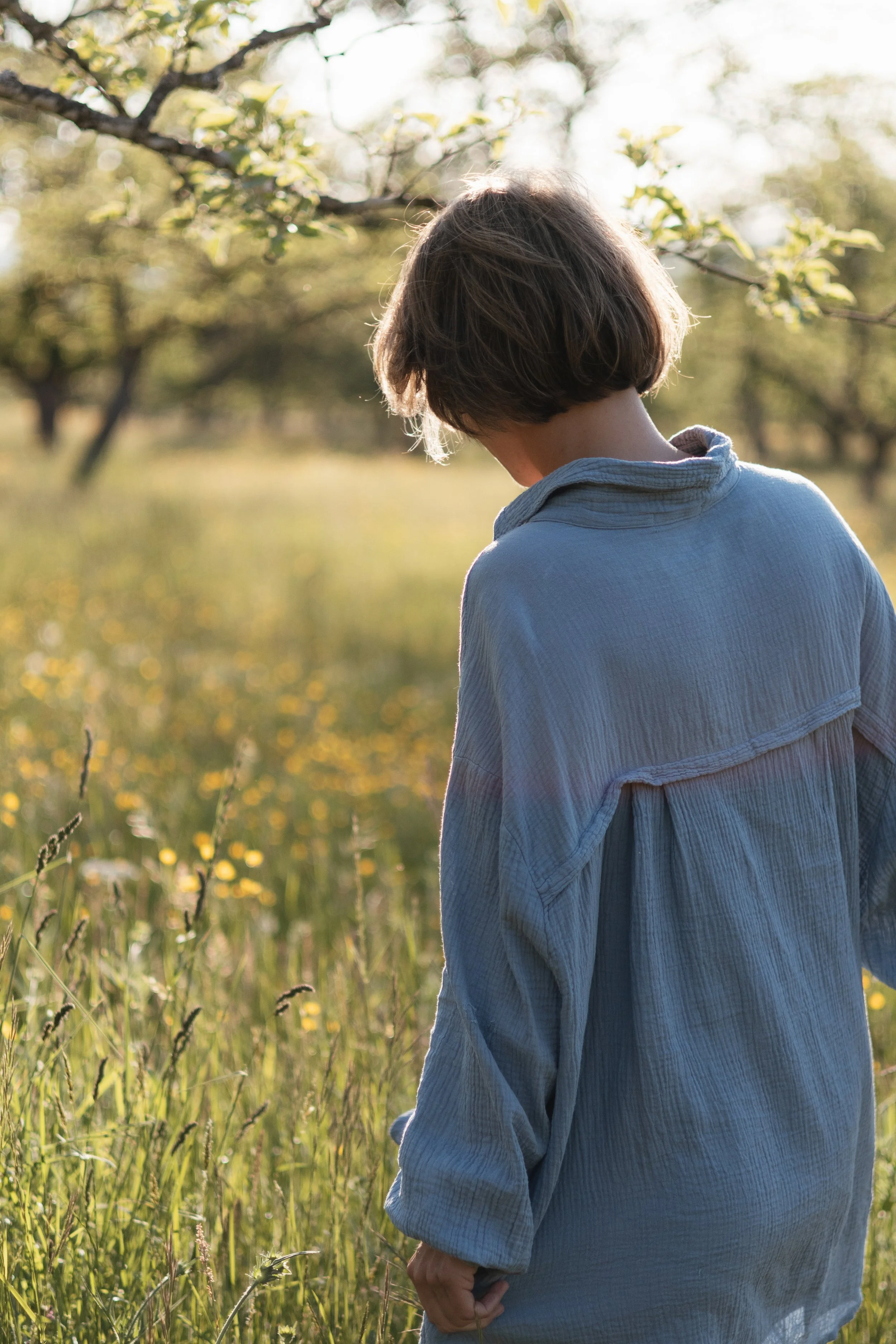 Person mit kurzen braunen Haaren, die nach hinten blickt, steht in einem Feld mit Blumen im Sonnenlicht, Bäumen im Hintergrund, trägt eine blaue Bluse.