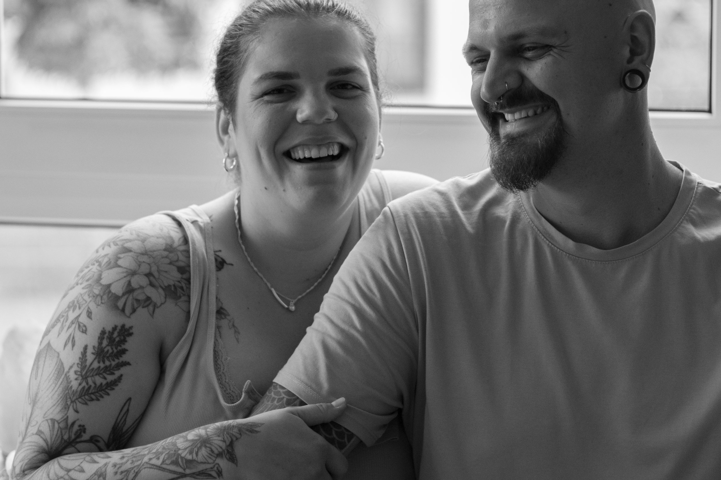 Zwei lachende Menschen sitzen nebeneinander vor einem Fenster, sie scheinen Freude zu teilen, bei einem ist die rechte Hand auf dem Arm des anderen.