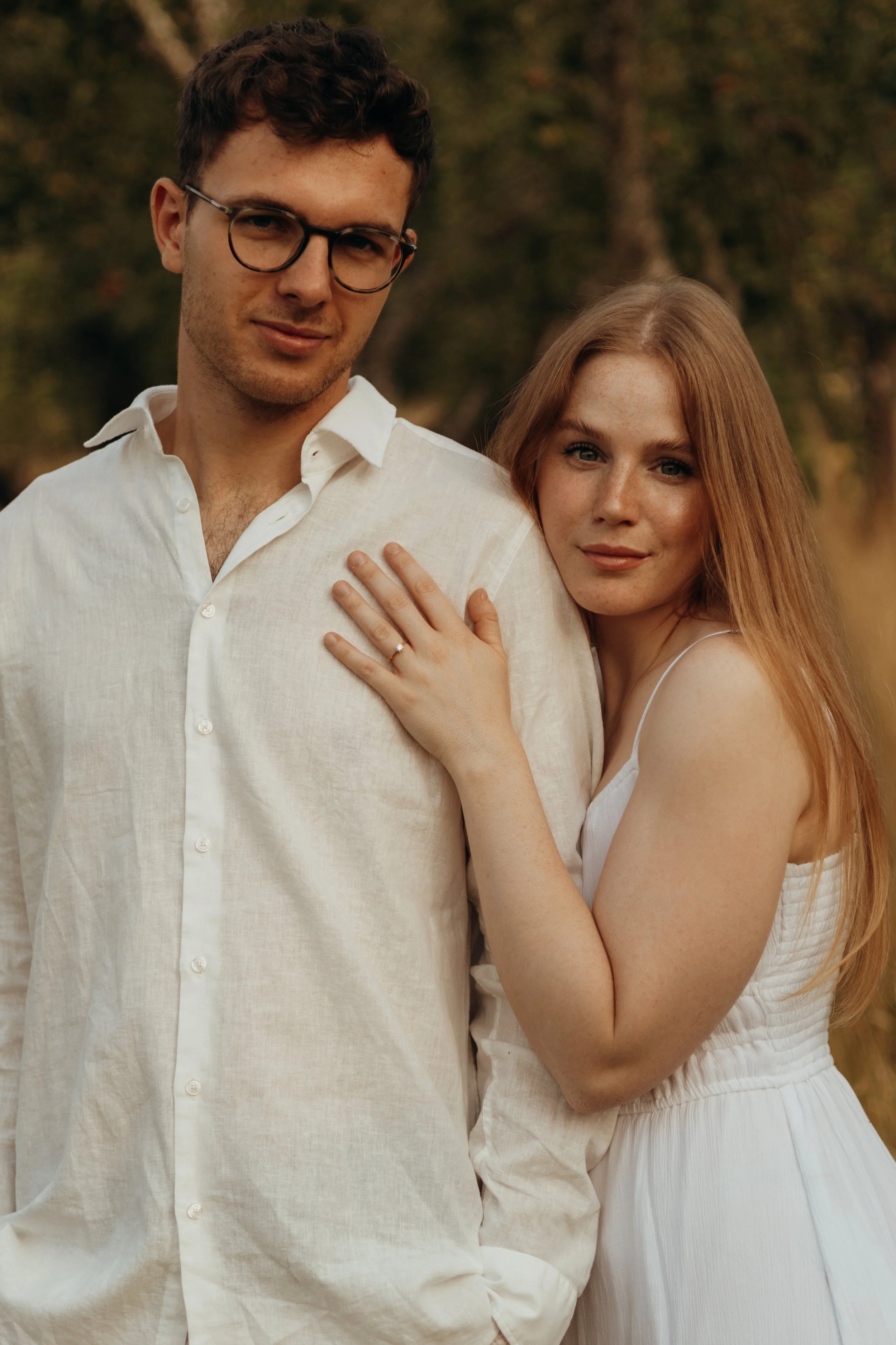 Ein junger Mann mit Brille und einer jungen Frau mit roten Haaren, die sich umarmen und auf einem Waldweg stehen.