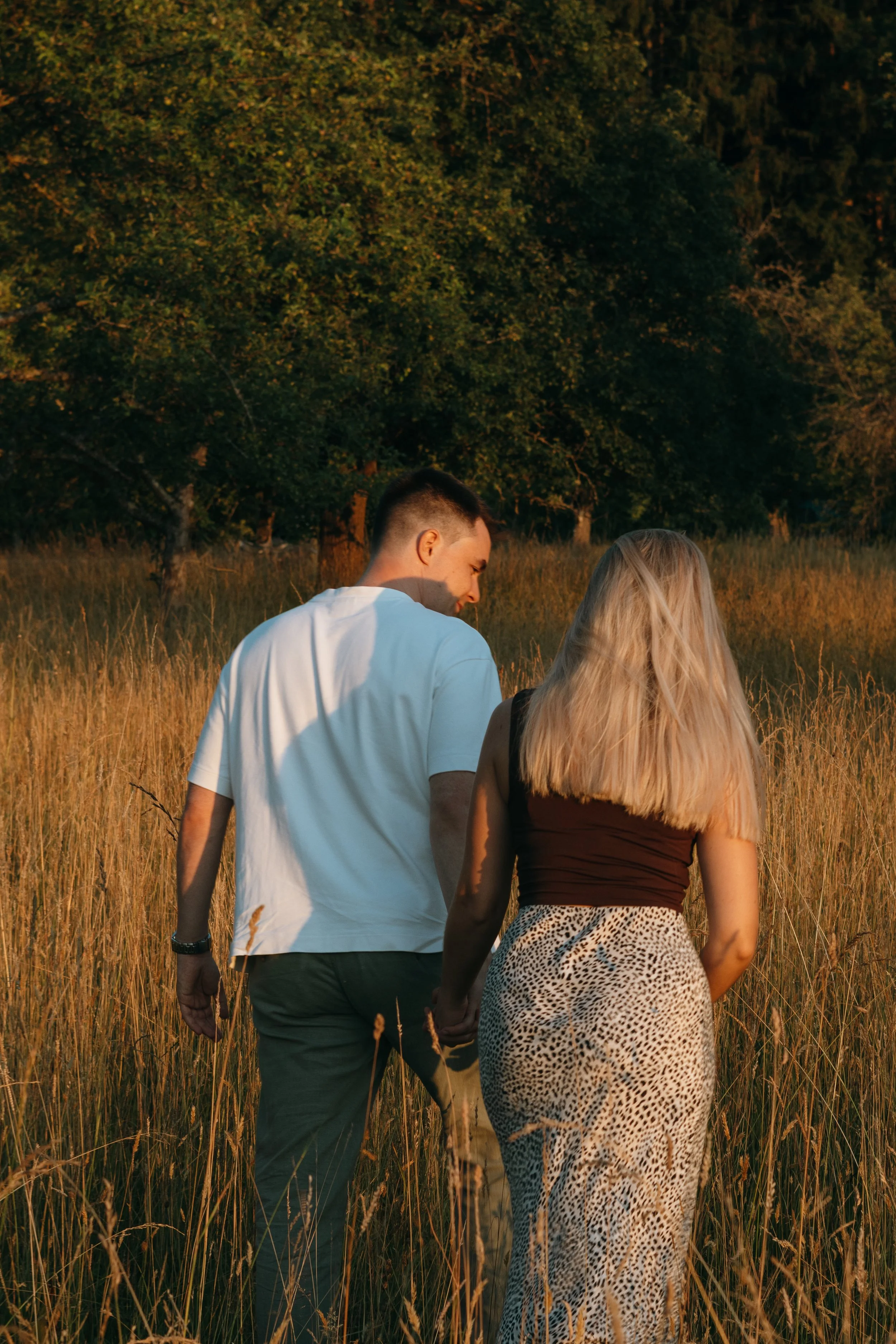 Ein Paar geht Hand in Hand durch ein sonnenverwöhntes Feld mit hohen Gräsern, umgeben von Bäumen bei Sonnenuntergang.