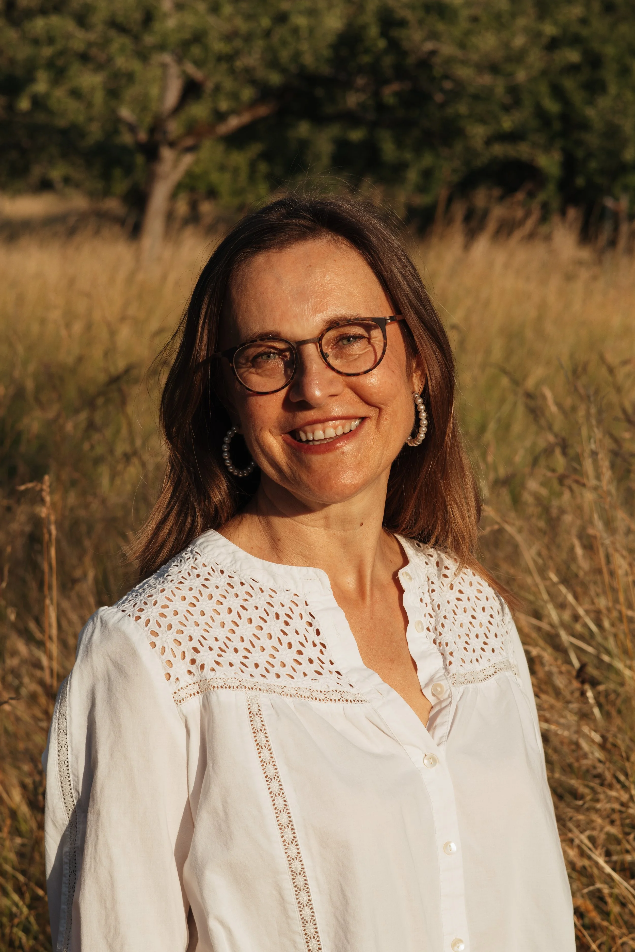 Ein lächelnder Frau mit Brille und Ohrringen, getragen in einem weißen, perforierten Top, im Freien bei Sonnenuntergang.