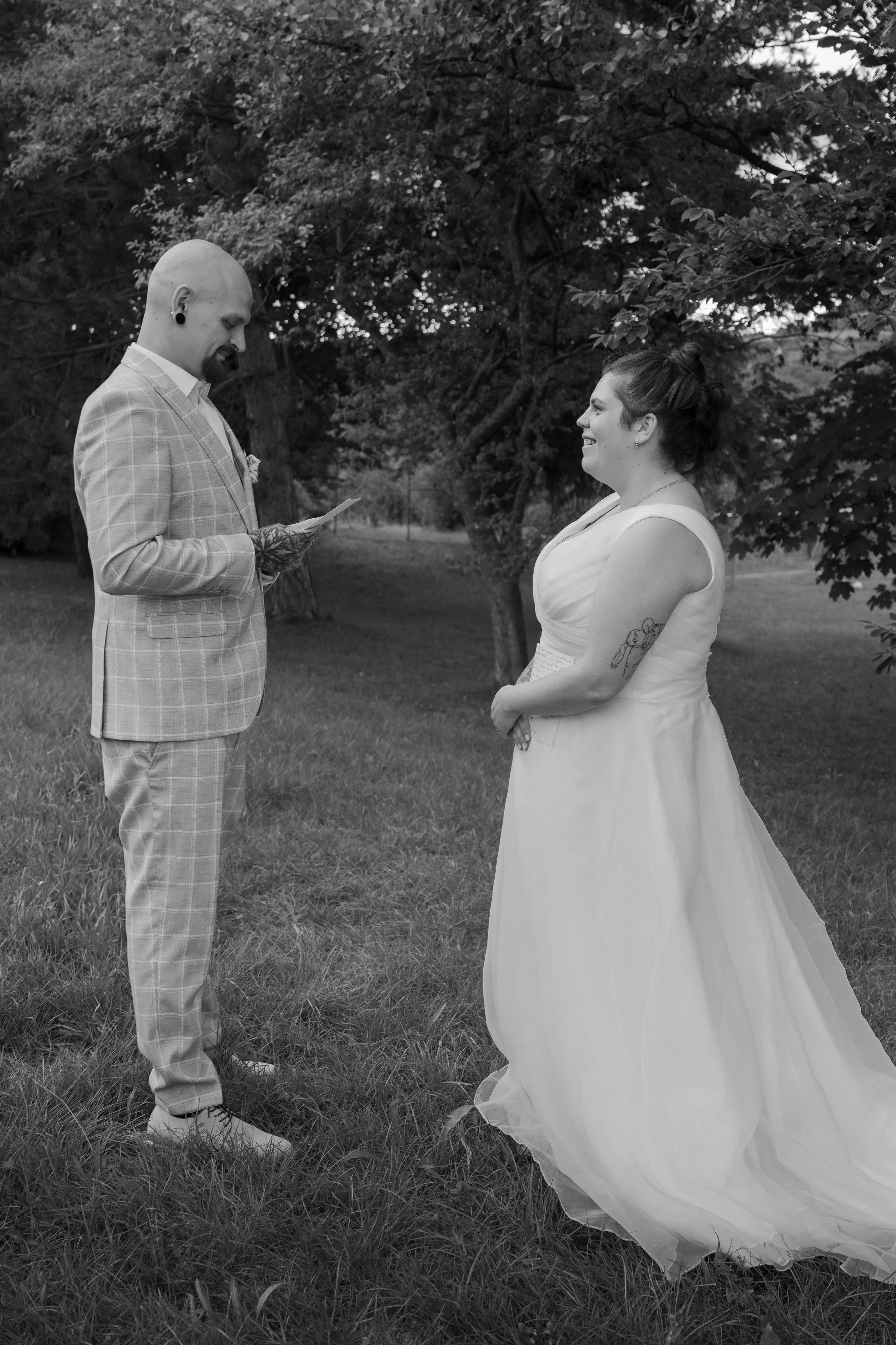 Ein Brautpaar bei einer standesamtlichen Trauung im Freien, die Braut trägt ein langes weißes Kleid und der Bräutigam ein kariertes Anzug. Beide lächeln sich an, es ist taghell und im Hintergrund sind Bäume.