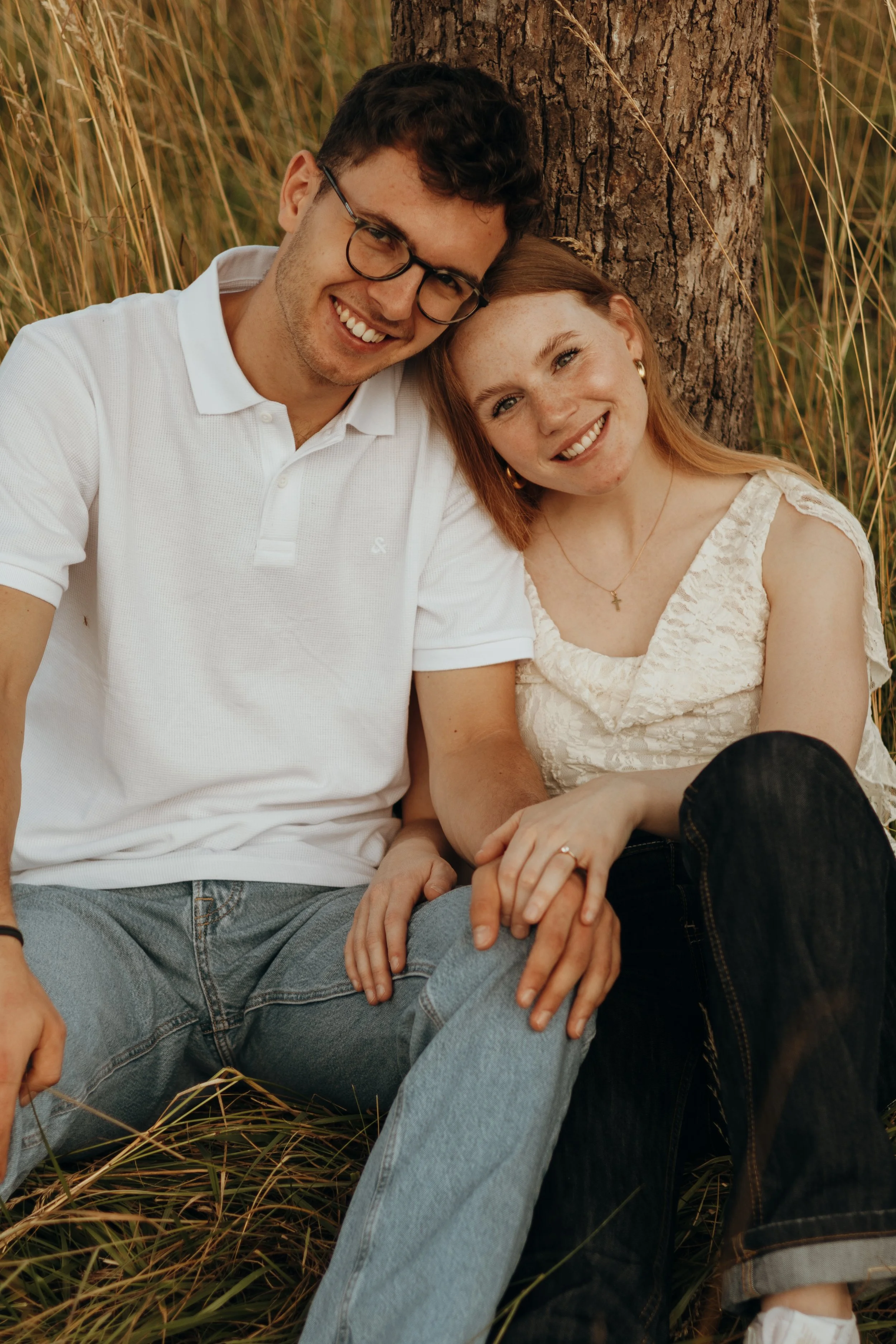 Ein glückliches junges Paar sitzt an einem Baum im Freien, umgeben von Gräsern, und lächelt in die Kamera.