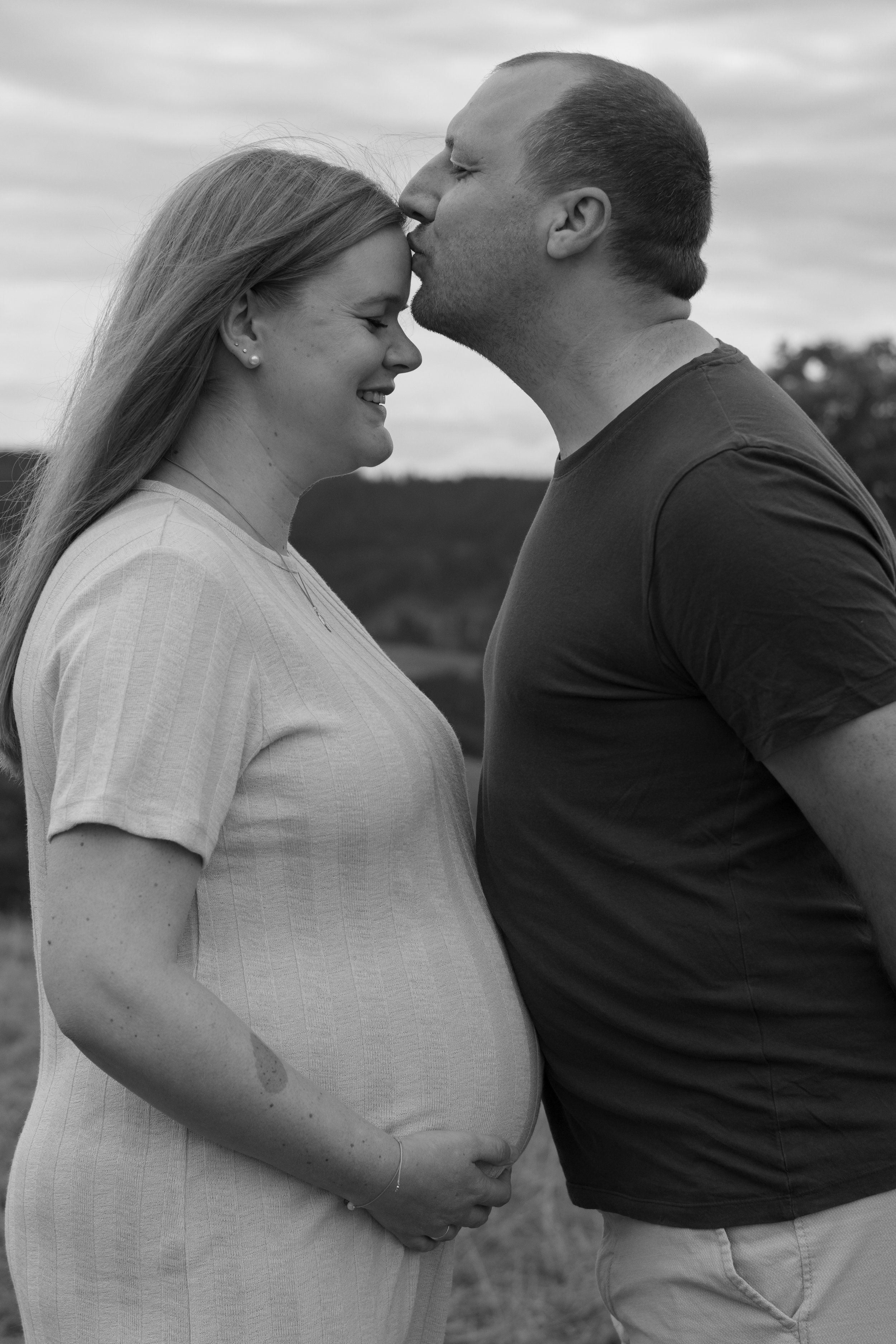 Ein schwangeres Paar in einer ländlichen Umgebung, der Mann küsst die Stirn der schwangeren Frau