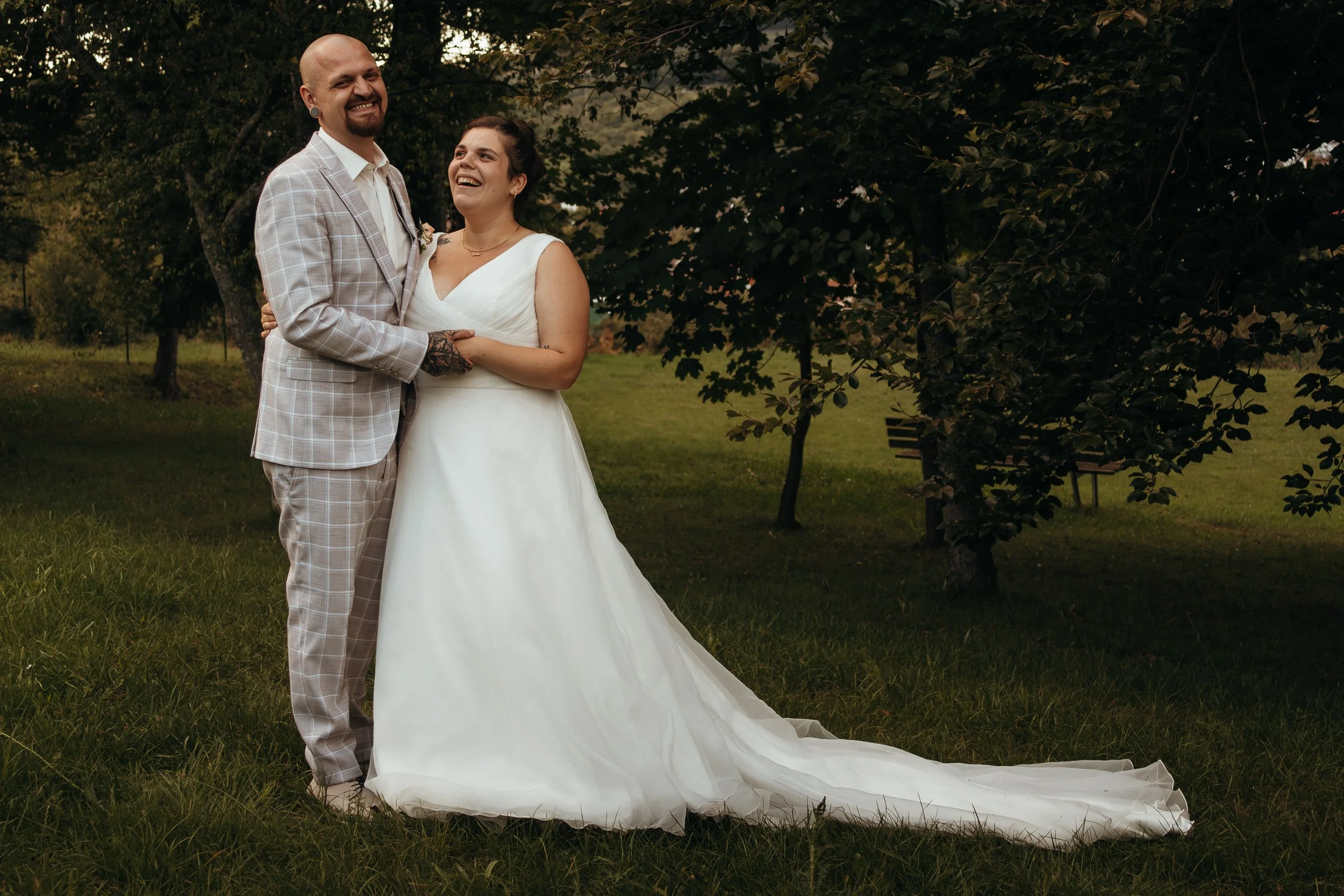 Ein lächelndes Brautpaar, das im Garten steht, die Braut in einem weißen Kleid, der Bräutigam in einem hellen Anzug.