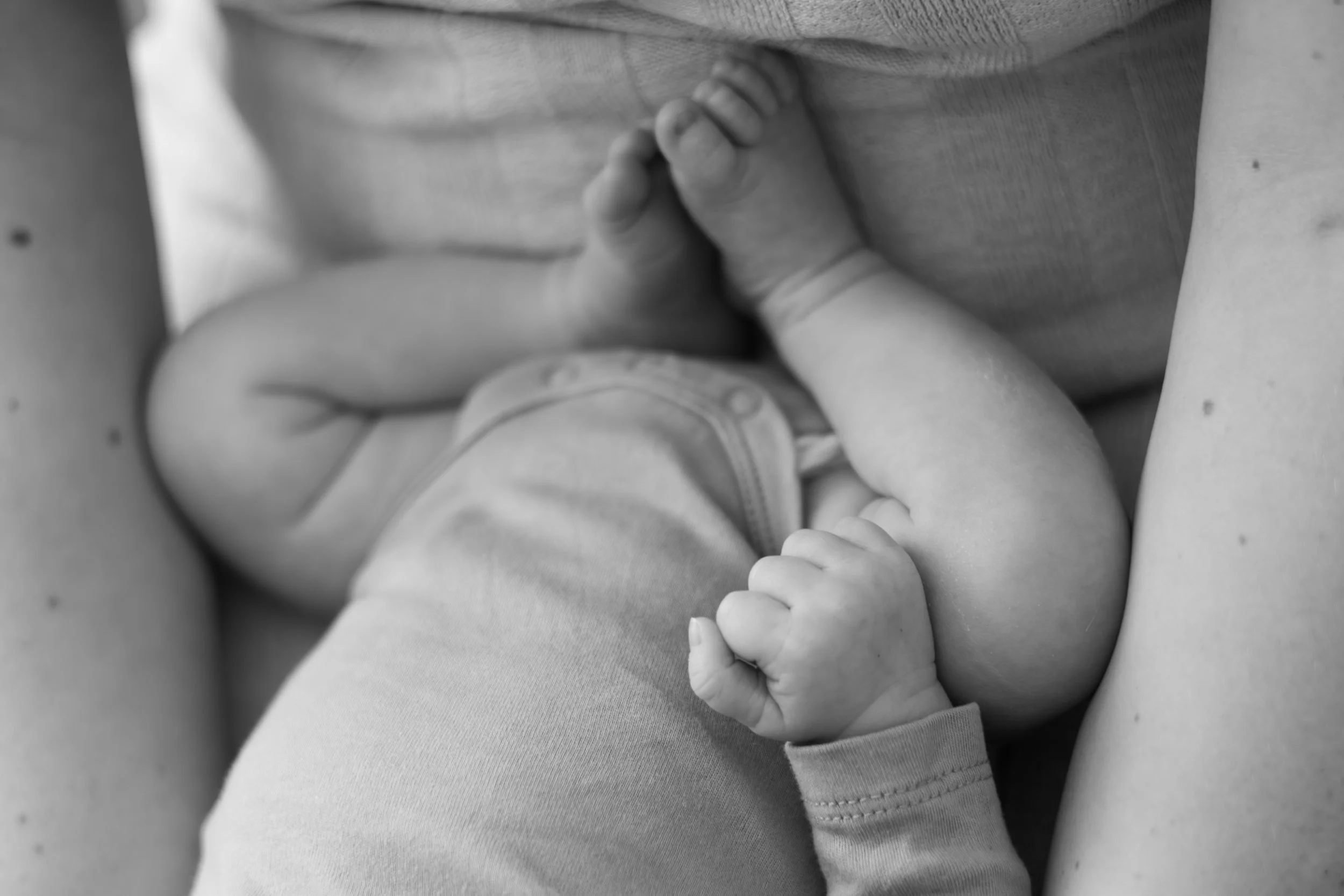 Schwarz-weiß-Bild eines Babys, das im Schoß einer Person liegt. Das Baby ist in einem onesie gekleidet und hat eine geballte Faust, sichtbar nur die Hand und der Arm sind, da der Rest des Babys outsichtbar ist.