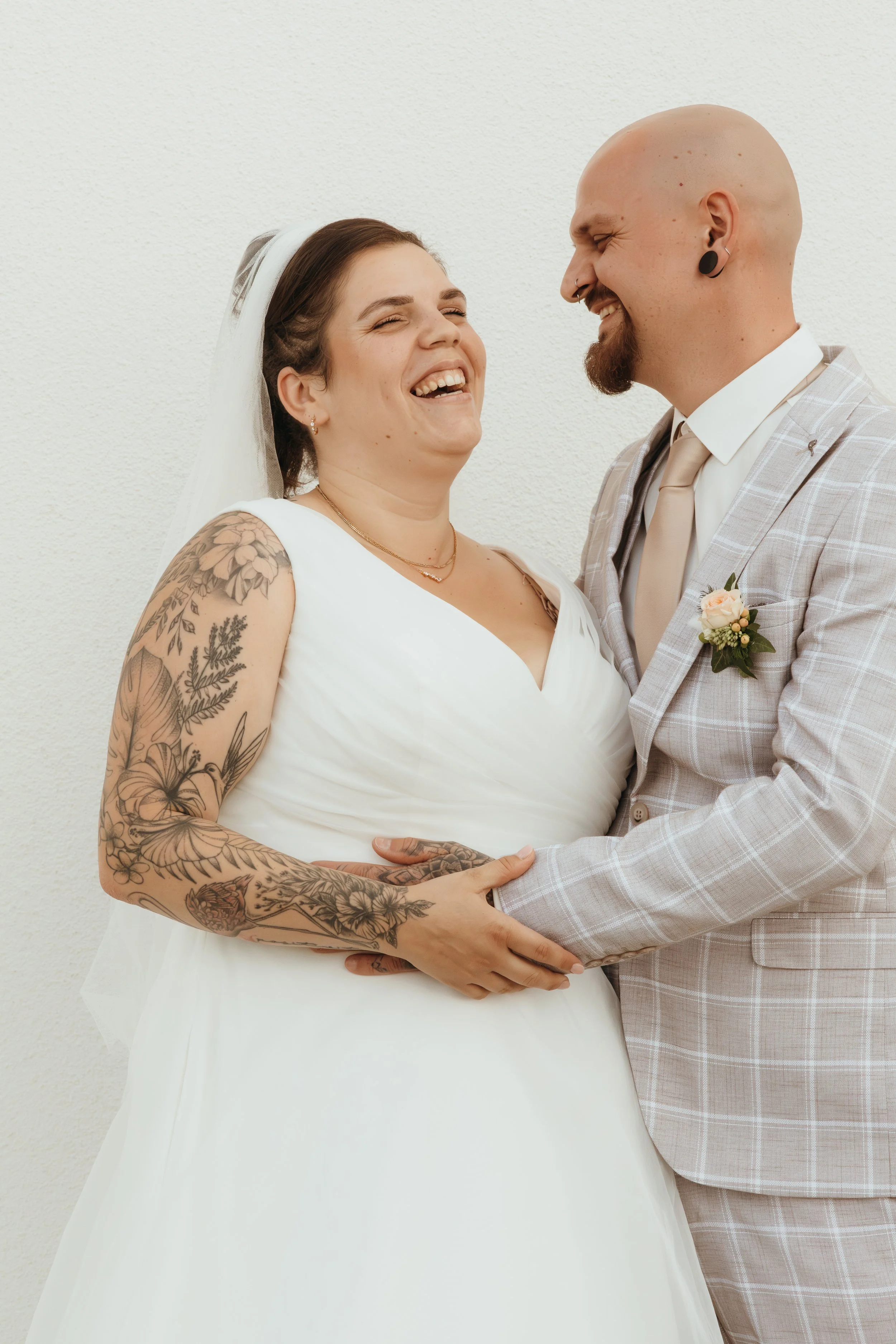 Ein lachendes lesbisches Hochzeitspaar, ein Mann in einem grau karierten Anzug und eine Frau in einem weißen Brautkleid mit Tattoos, hält sich an den Händen, vor einer weißen Wand.