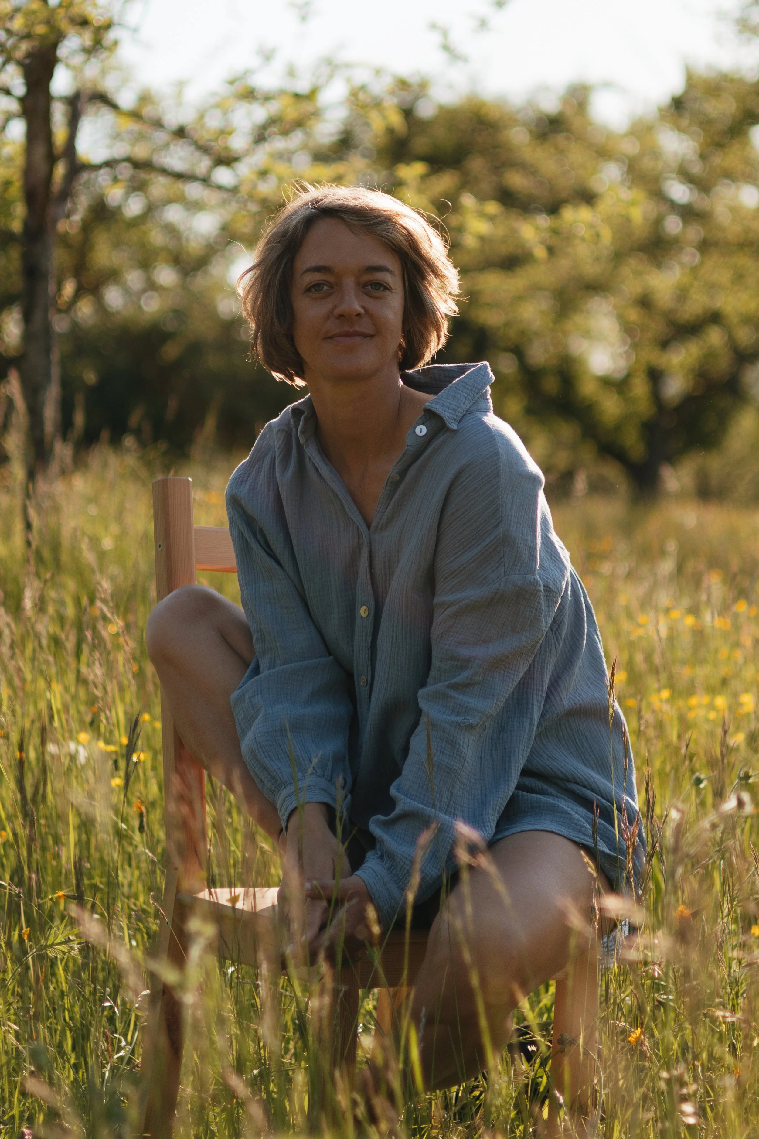 Frau sitzt auf einem Holzstuhl in einem Feld mit hohen Gräsern und kleinen gelben Blumen, bei Sonnenuntergang, im Ruhezustand, trägt eine blaue Hemdbluse.