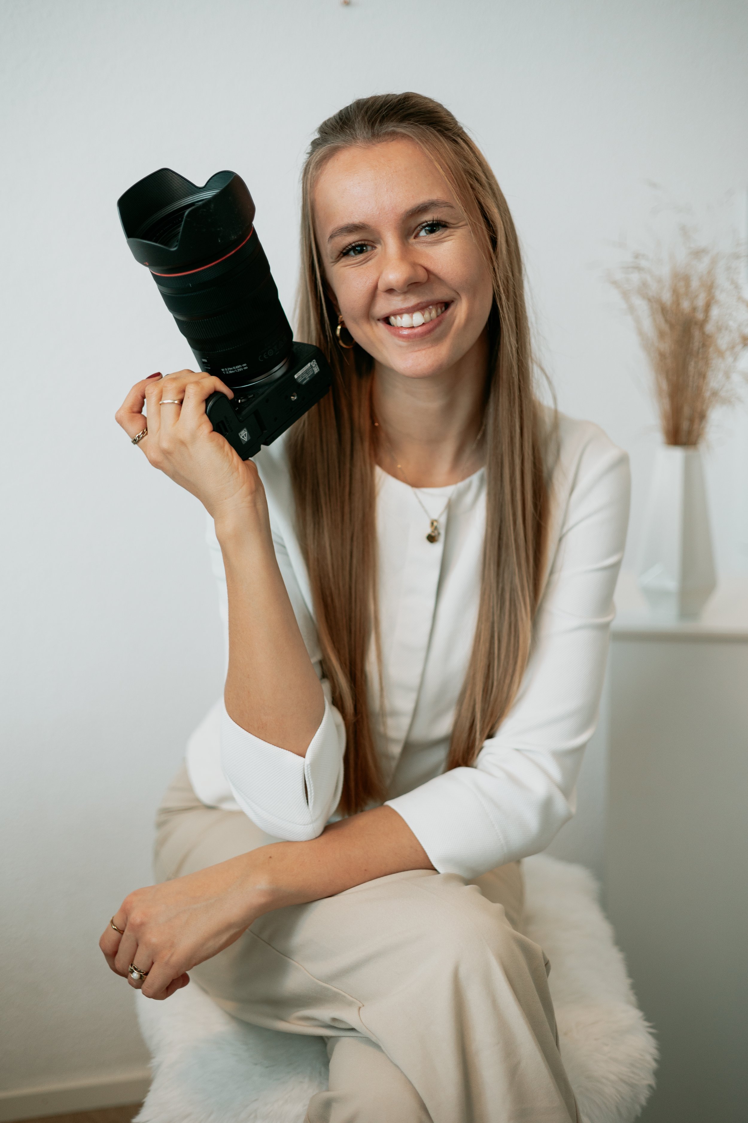 Eine lachende junge Frau mit langem braunem Haar hält eine Kamera in der Hand und sitzt auf einem weißen Hocker mit einem Fellbezug. Im Hintergrund sind getrocknete Pflanzen in einer weißen Vase sichtbar.
