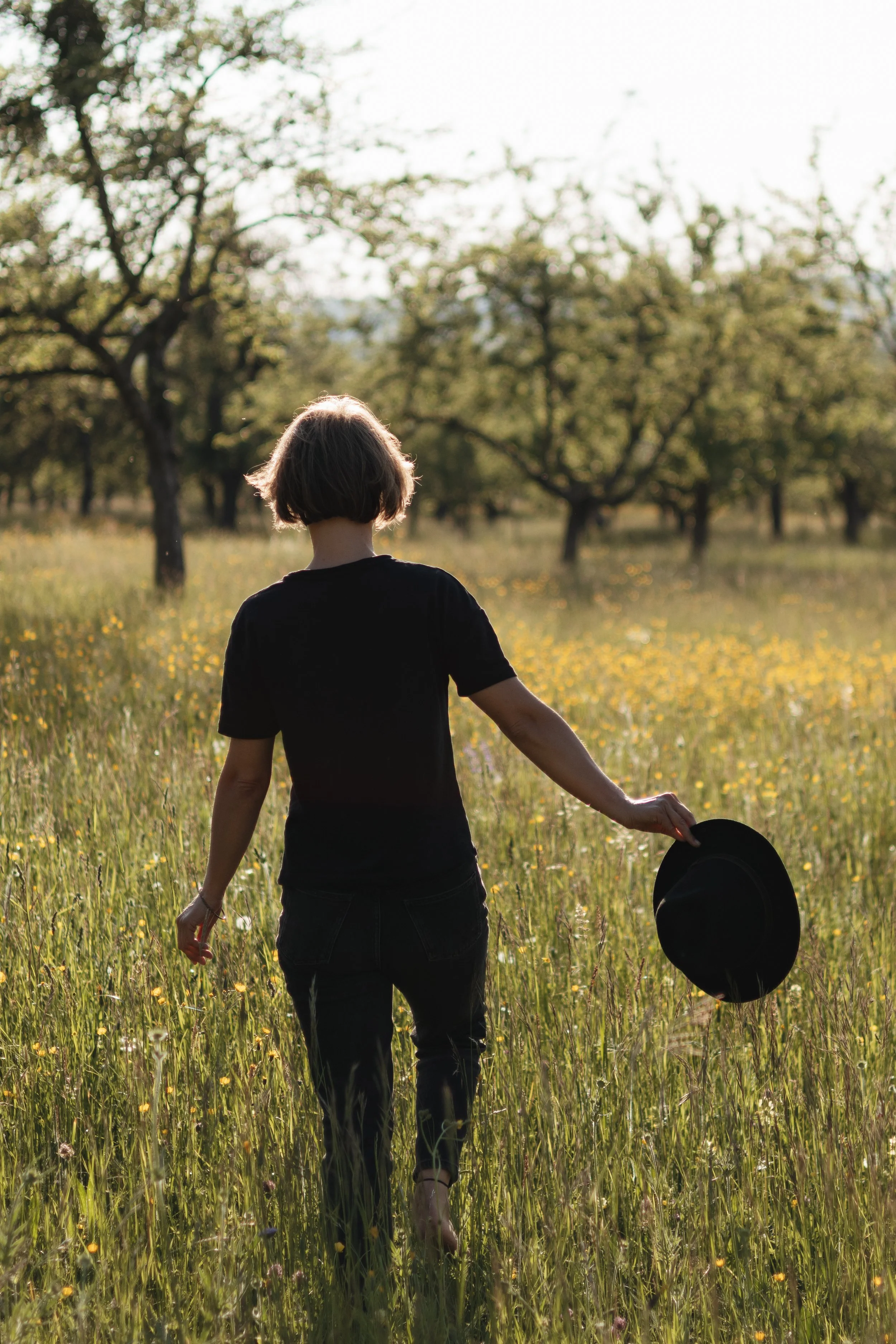 Eine Person mit kurzem Haar, schwarze Kleidung und Hut in einem blühenden Feld bei Sonnenuntergang, von hinten fotografiert.