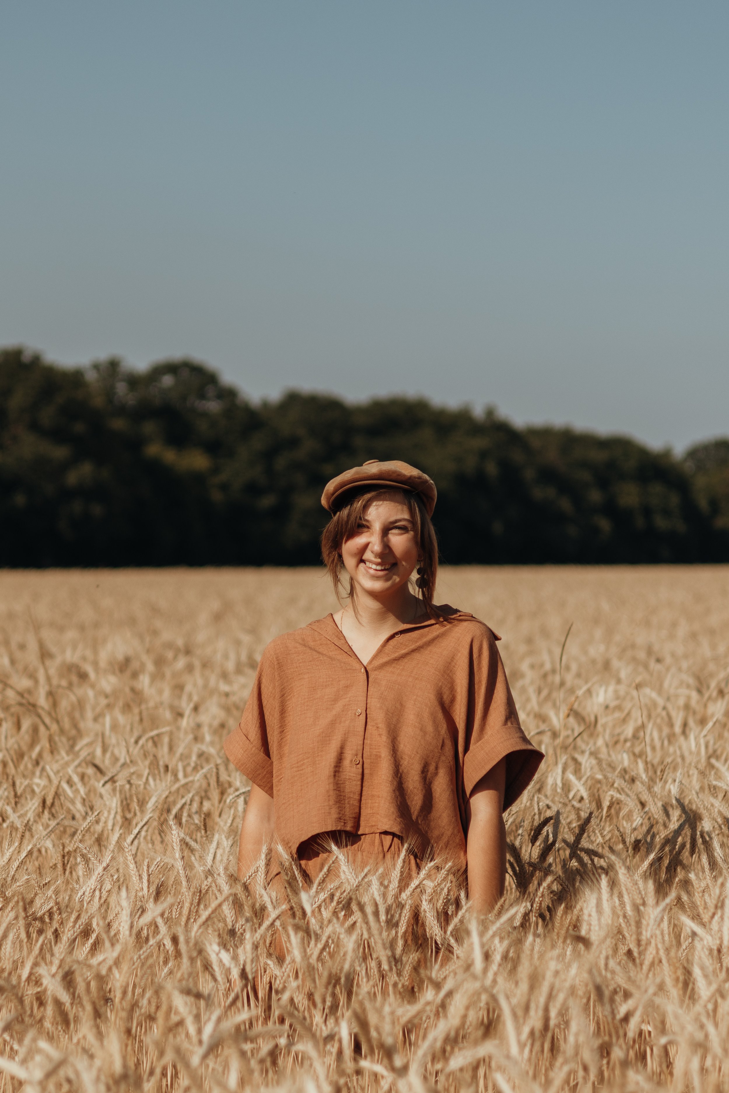 Eine lachende Frau trägt eine braune Jacke und einen Hut, steht in einem goldgelben Weizenfeld bei Sonnenschein, im Hintergrund Bäume und blauer Himmel.