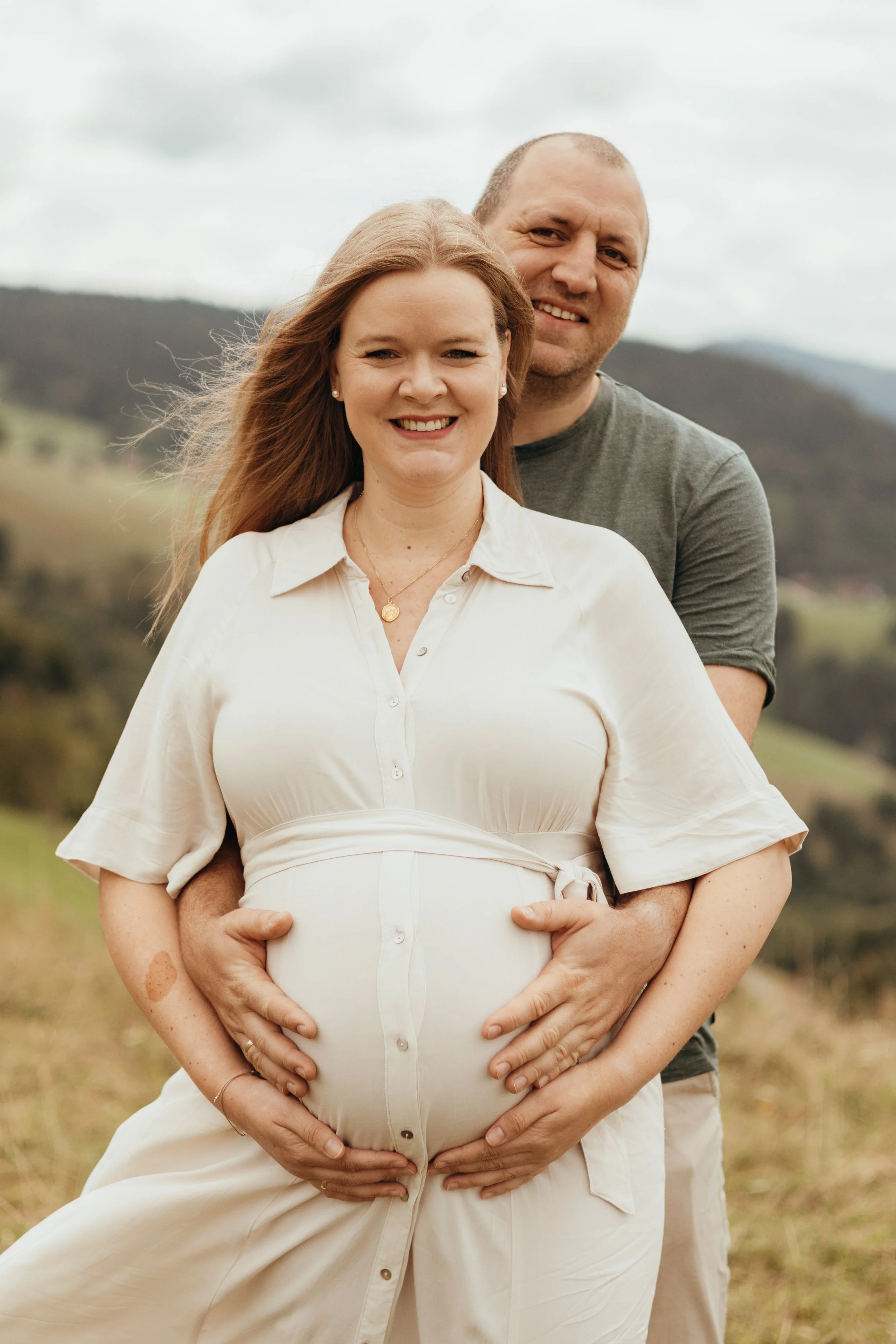Ein glückliches schwangere Paar, das draußen in einer ländlichen Umgebung steht, mit den Händen auf dem Babybauch, bei einem Fotoshooting.