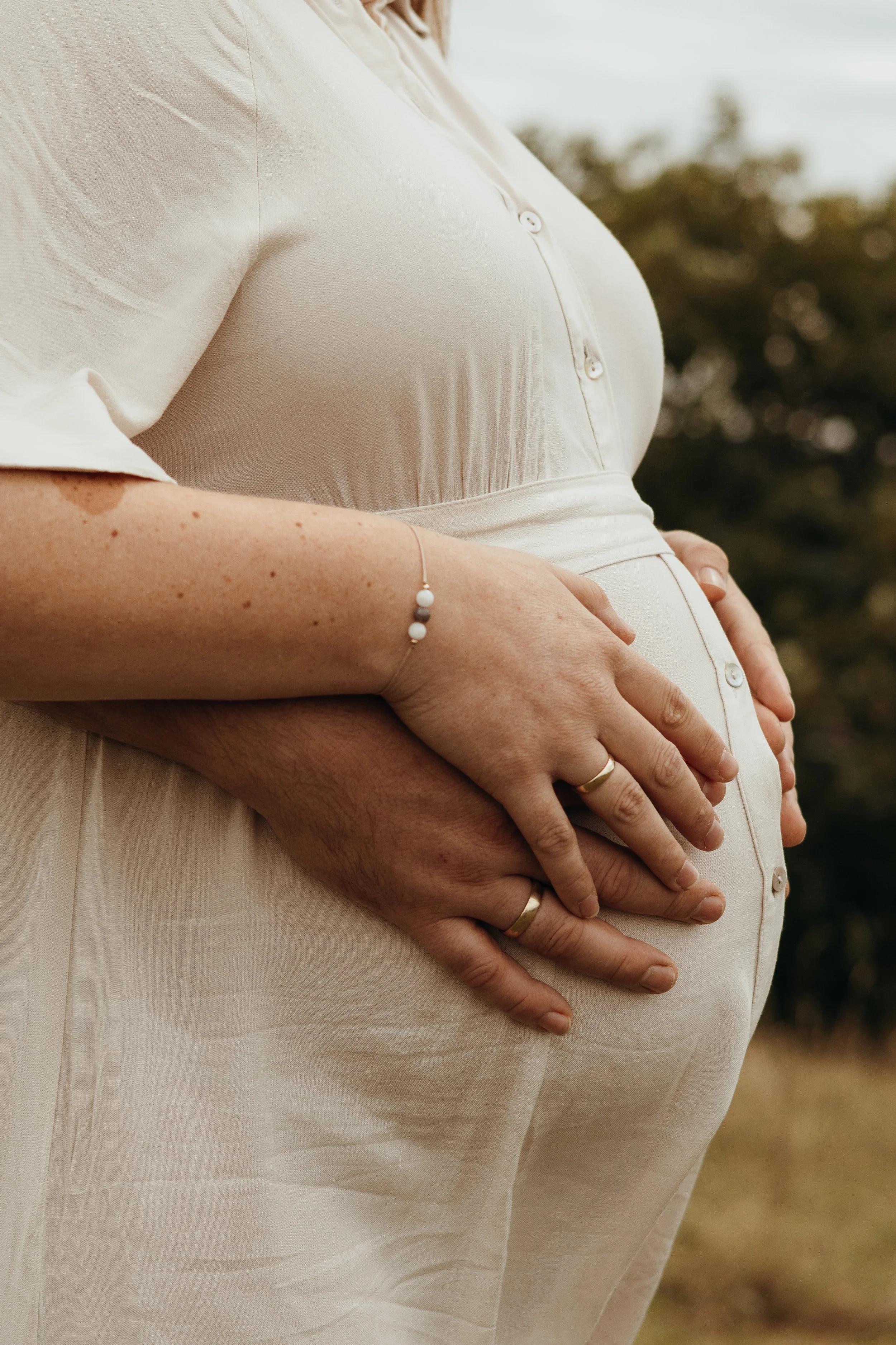 Nahaufnahme eines schwangeren Paares, das seine Hände auf den Bauch legt, draußen im Freien bei bewölktem Himmel.