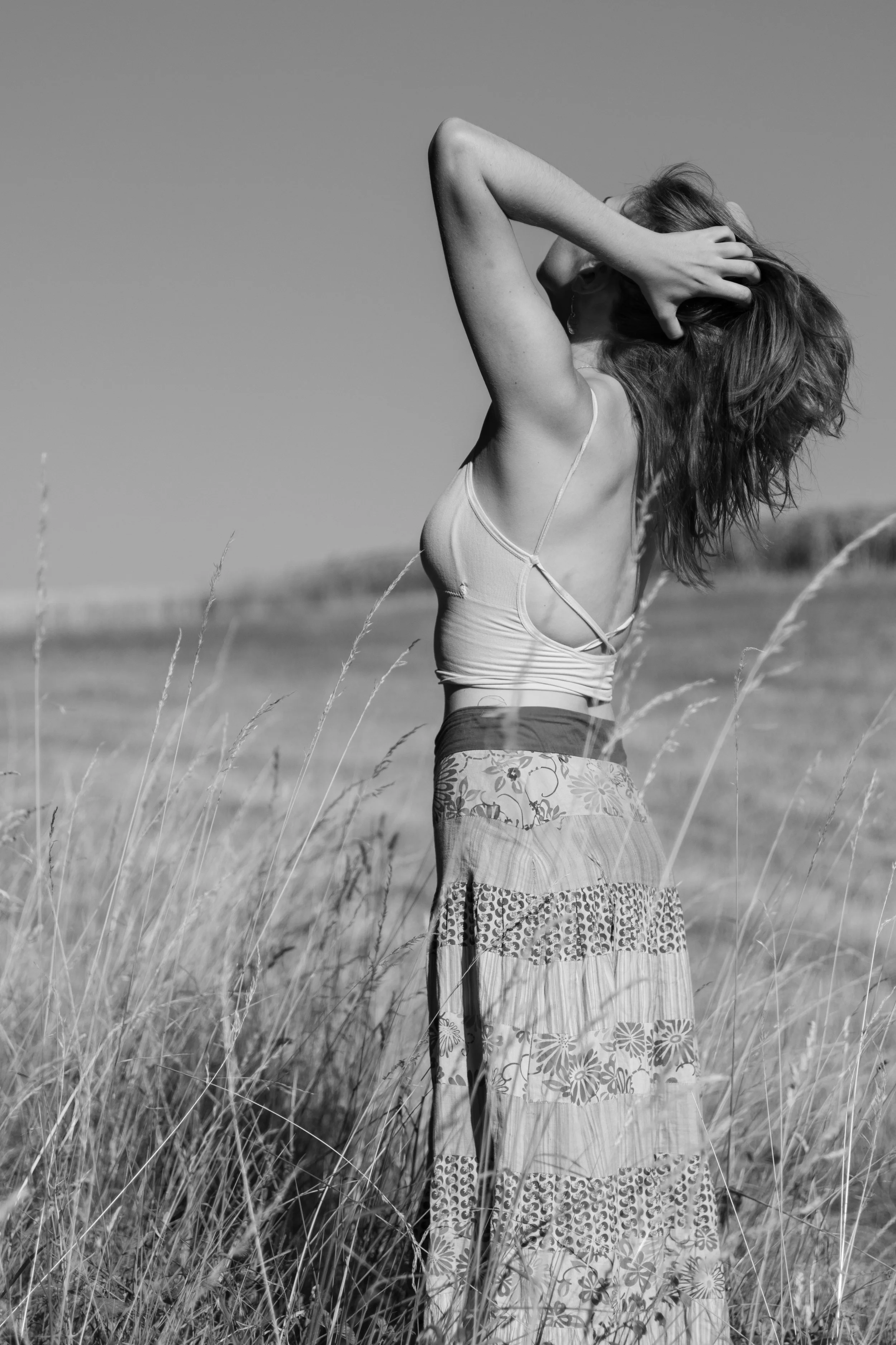 Frau steht in einem Feld aus hohem Gras, hebt ihre Arme und berührt ihren Kopf, trägt ein Top und einen langen, bunten Rock, im Schwarz-Weiß-Foto.