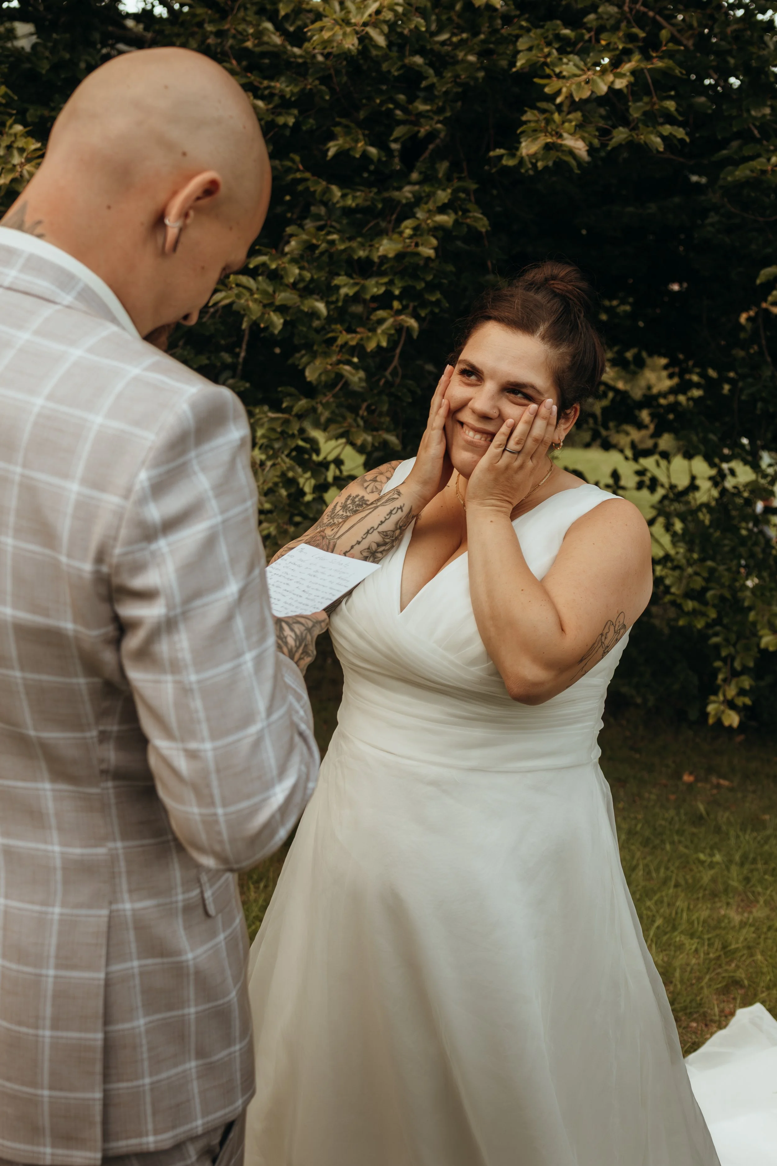 Ein Mann liest eine Handgeschriebene Rede vor einer Frau in einem weißen Hochzeitskleid. Das Paar ist im Freien vor einem Busch. Die Frau lächelt glücklich mit den Händen im Gesicht.