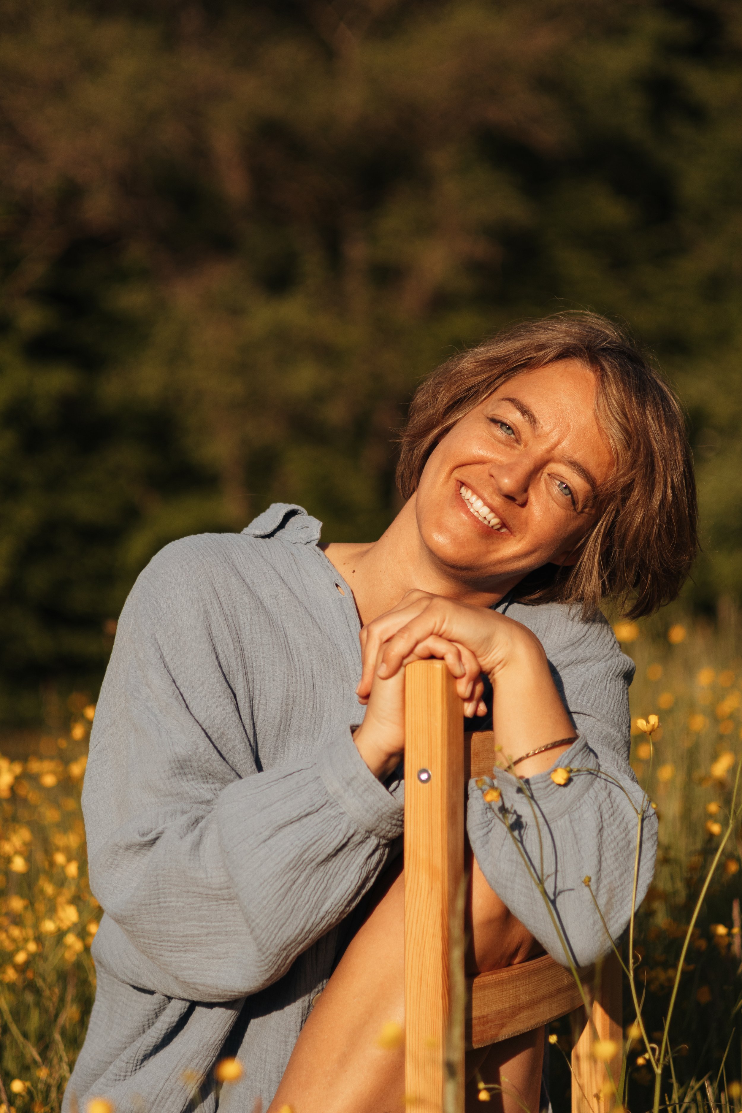Lächelnde Frau mit braunen Haaren, die im Sonnenlicht in einer Blumenwiese sitzt, die Arme auf einem Stuhl ruhend.