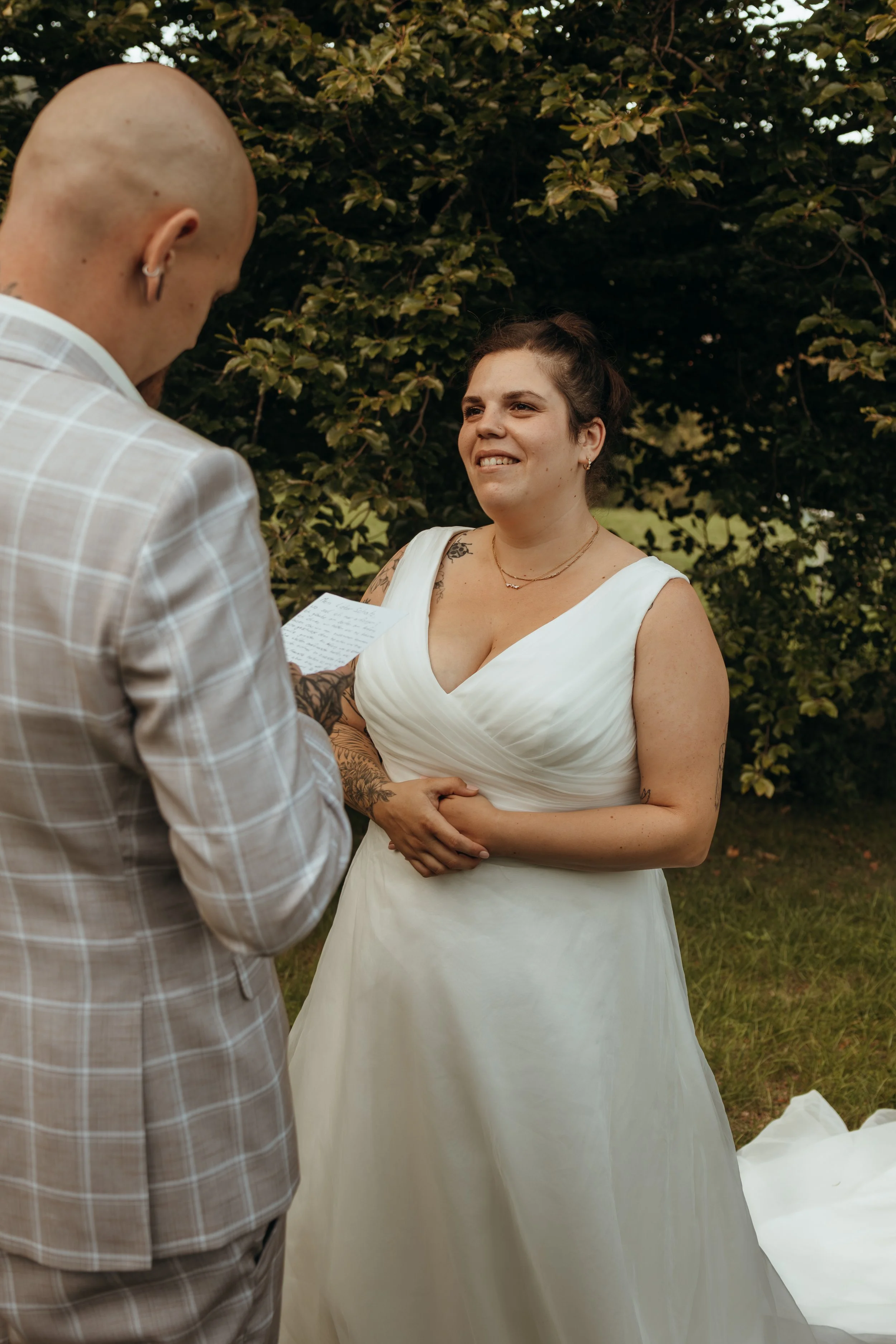 Eine brautähnliche Frau mit Tattoos in einem weißen Brautkleid beim Ja-Wort. Sie steht vor einem Mann, der eine Note oder einen Zettel hält. Sie befinden sich im Freien vor einem Laubhintergrund.