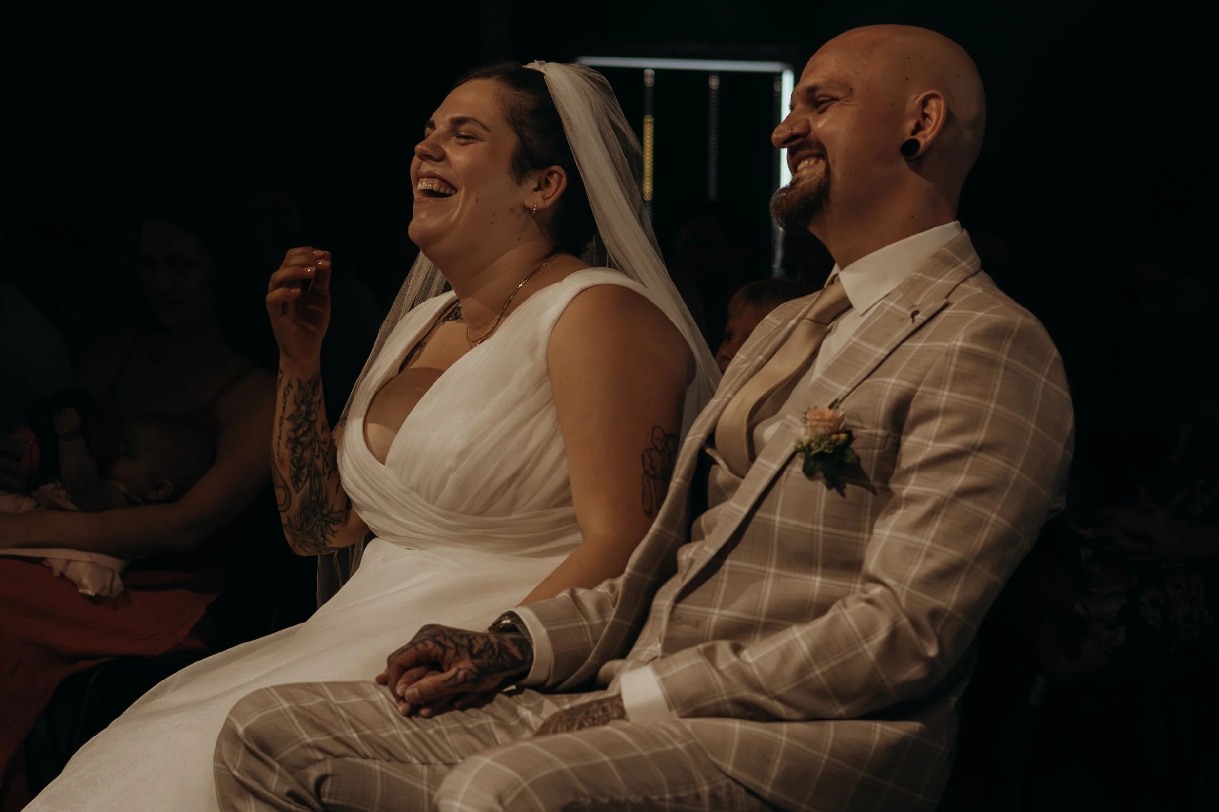 Ein Brautpaar sitzt lachend während einer Hochzeitszeremonie, die Braut trägt ein weißes Kleid und einen Schleier, der Bräutigam trägt einen beige Karoanzug mit einem Blumenanstecker.