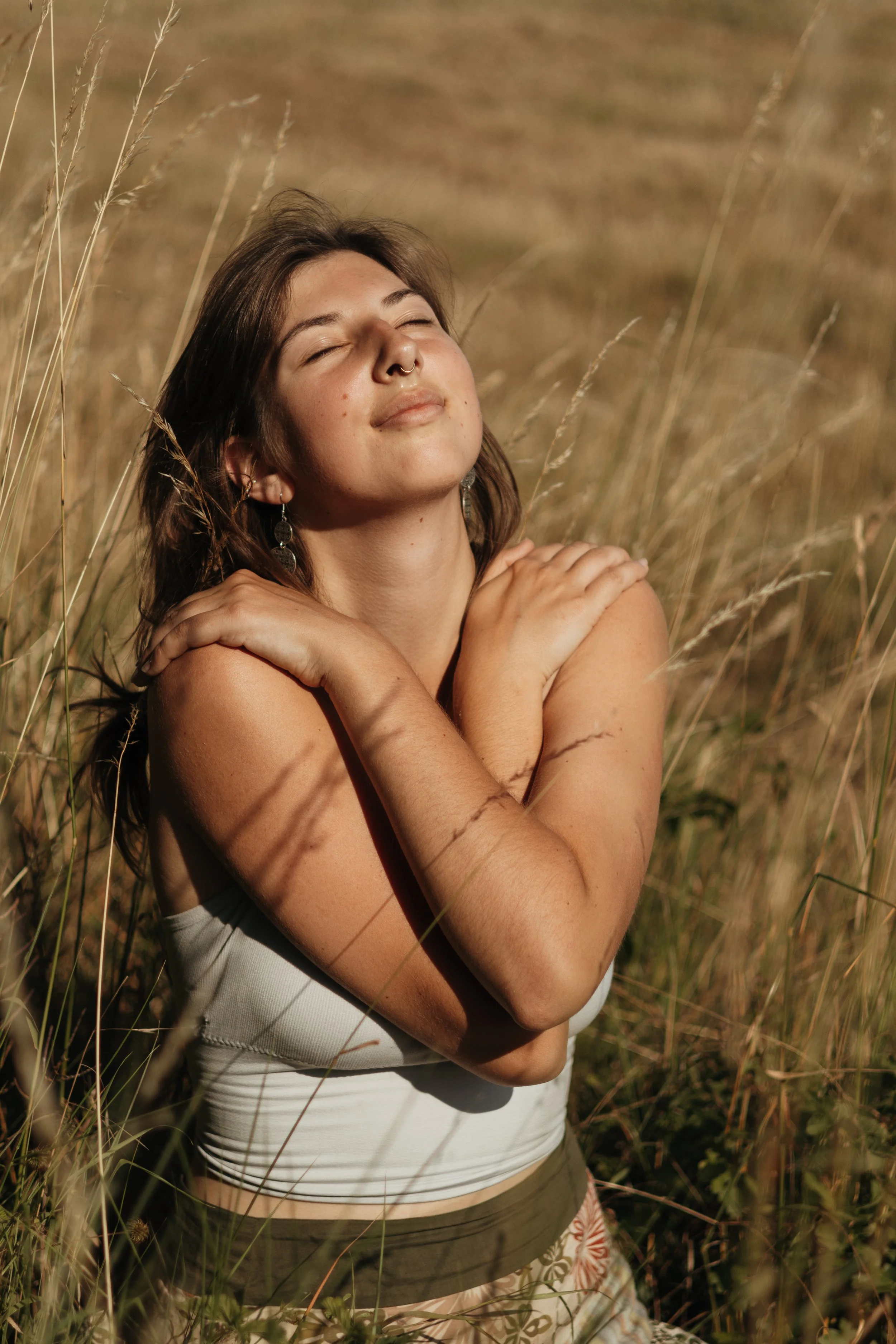 Eine junge Frau mit braunen Haaren, die die Augen geschlossen hat und die Hände auf die Schultern legt, steht in einem hohen Grasteppich im Sonnenlicht.
