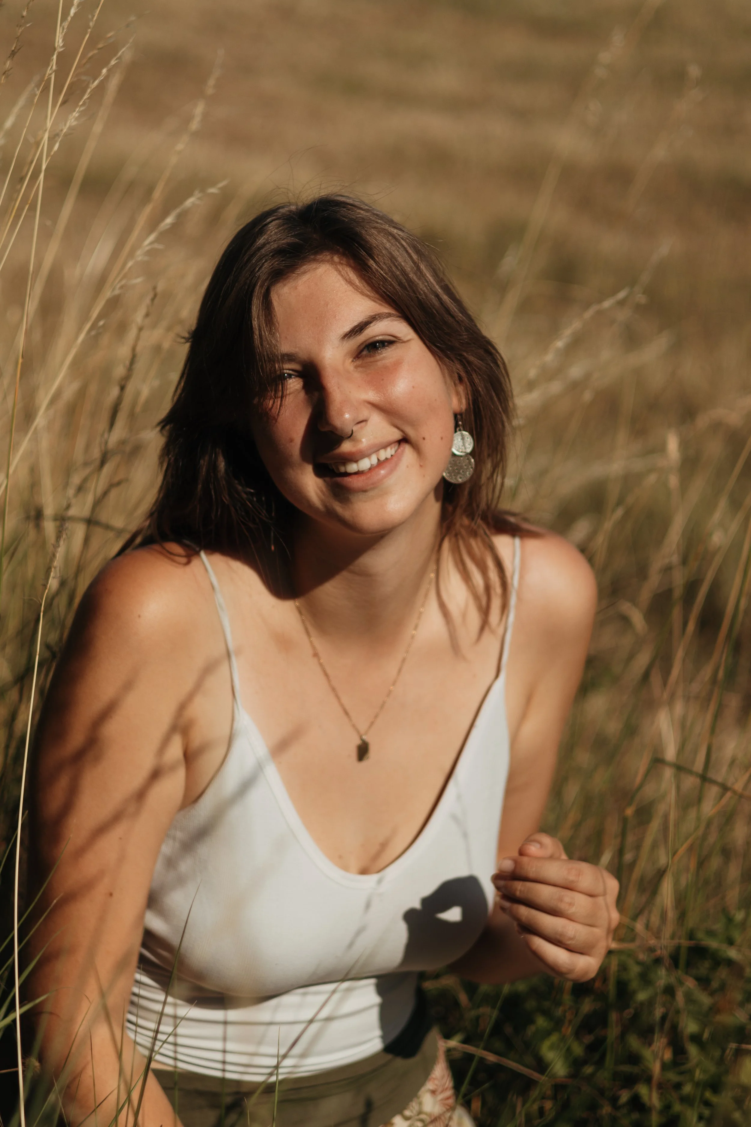 Lächelnde junge Frau mit braunen Haaren, weißen Top und Ohrringen im Sonnenlicht in einem Feld aus trockenem Gras.