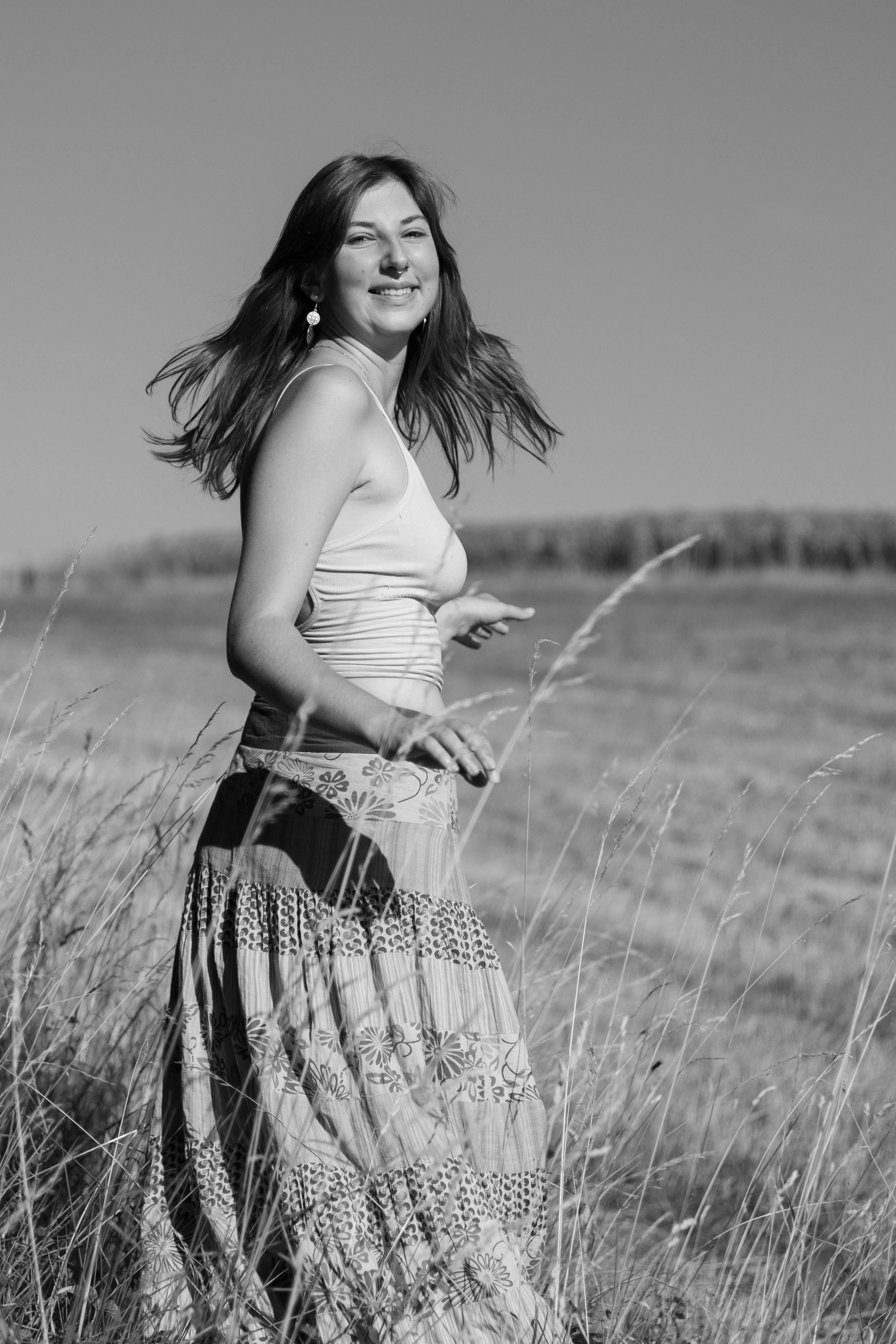 Eine junge Frau läuft durch eine Wiese an einem sonnigen Tag, trägt ein Tanktop und einen lang bedruckten Rock, lächelt und hat lange волосы.