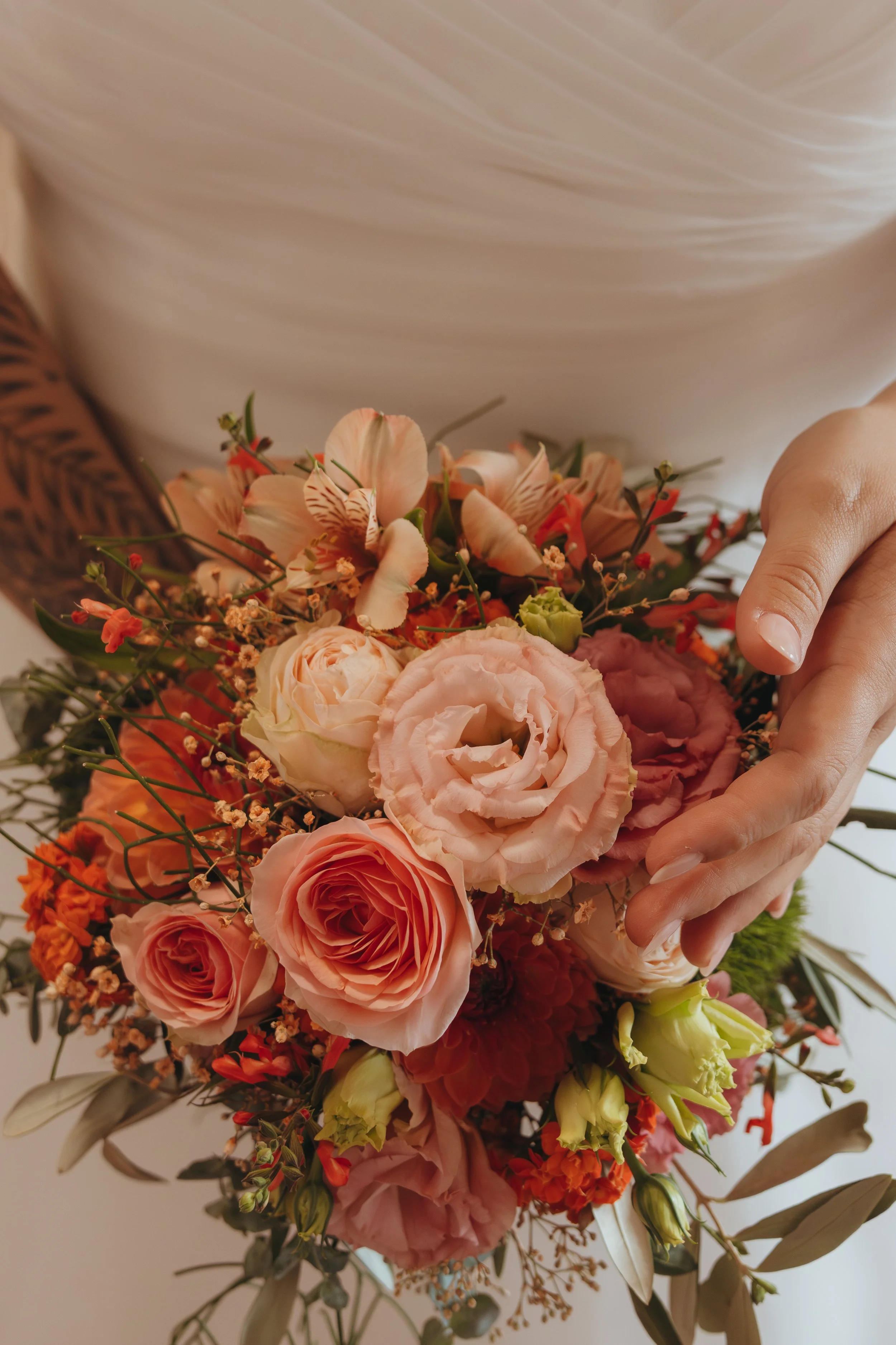 Eine Person hält einen Blumenstrauß mit rosa, weißen und orangefarbenen Blumen, darunter Rosen und andere Blüten. Der Hintergrund ist unscharf.