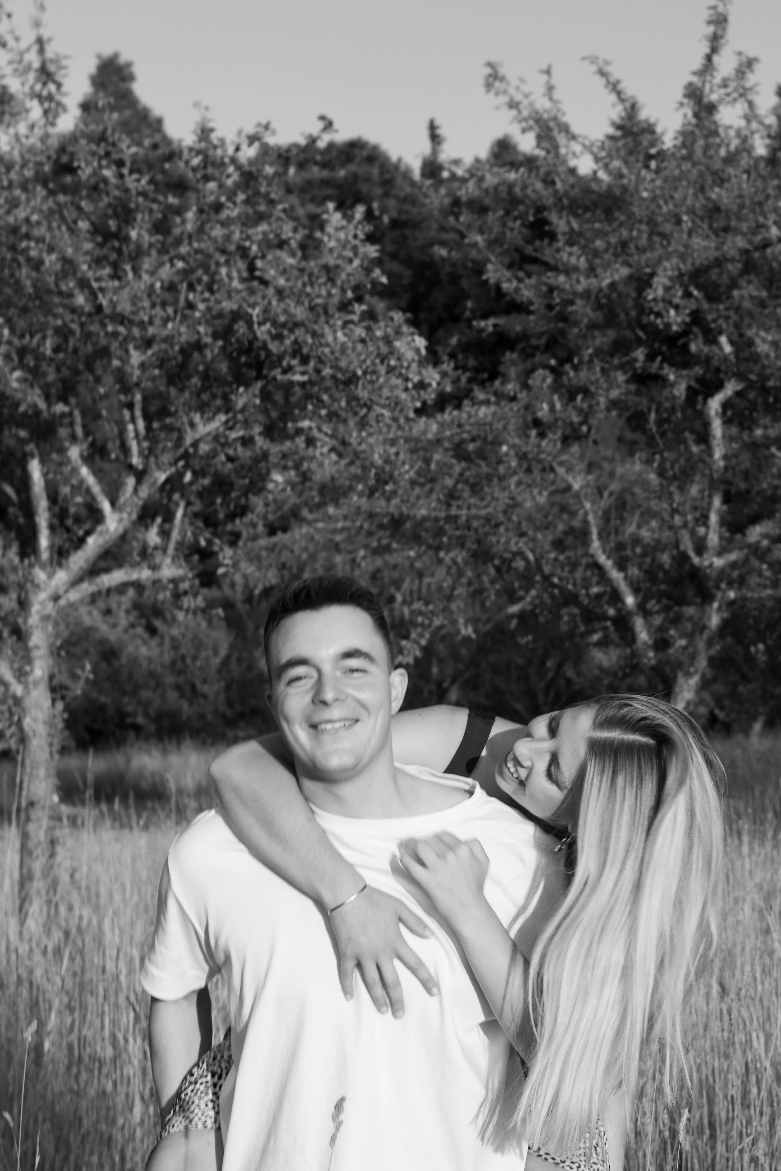 Ein lachendes Paar, das im Freien in einem Grasfeld mit Bäumen im Hintergrund eine Umarmung teilt, schwarz-weiße Aufnahme.