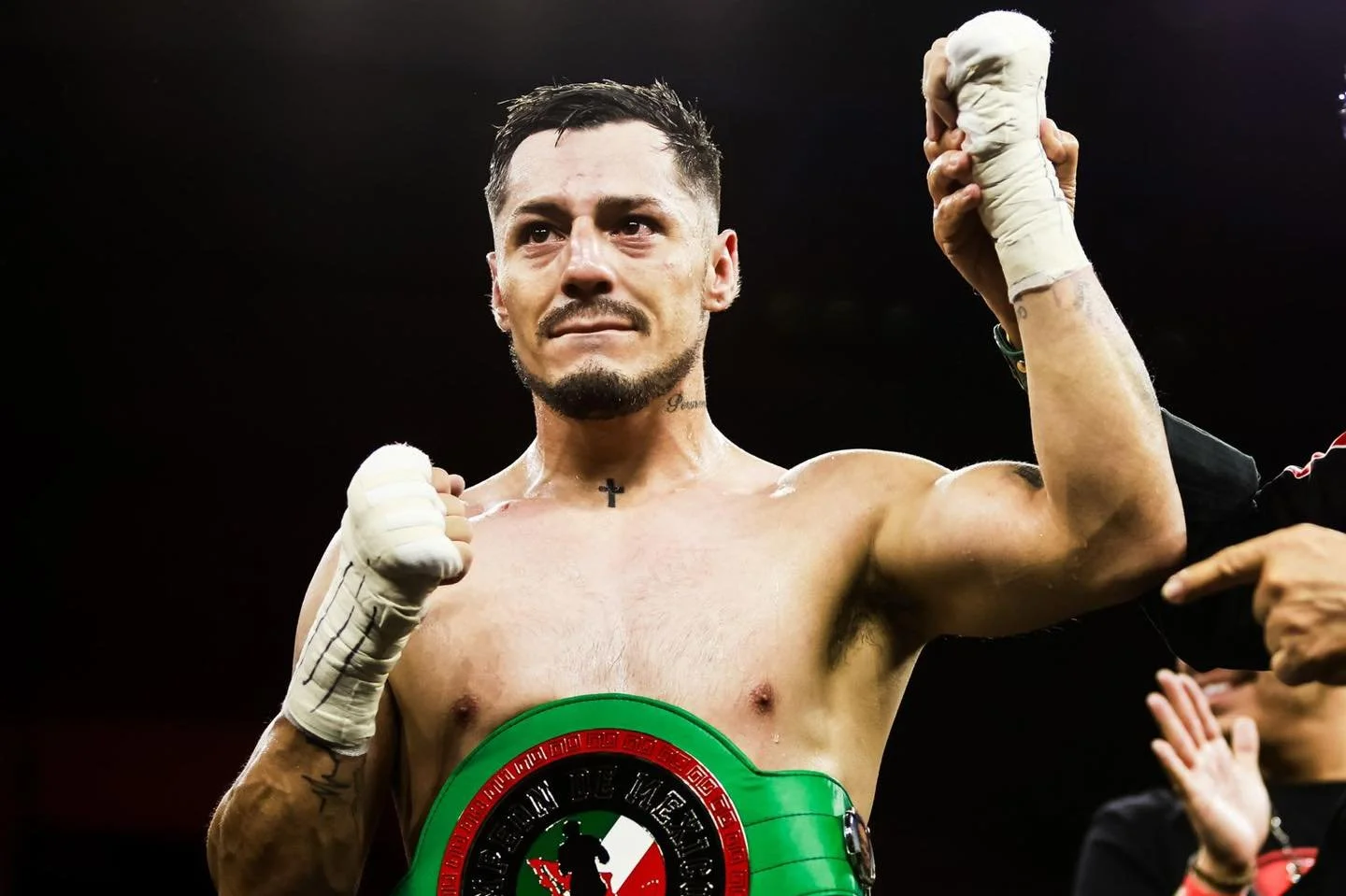 A shirtless male boxer with tape around his hands and a championship belt around his waist, flexing his right arm in a victory pose with a serious expression, standing in a boxing ring.