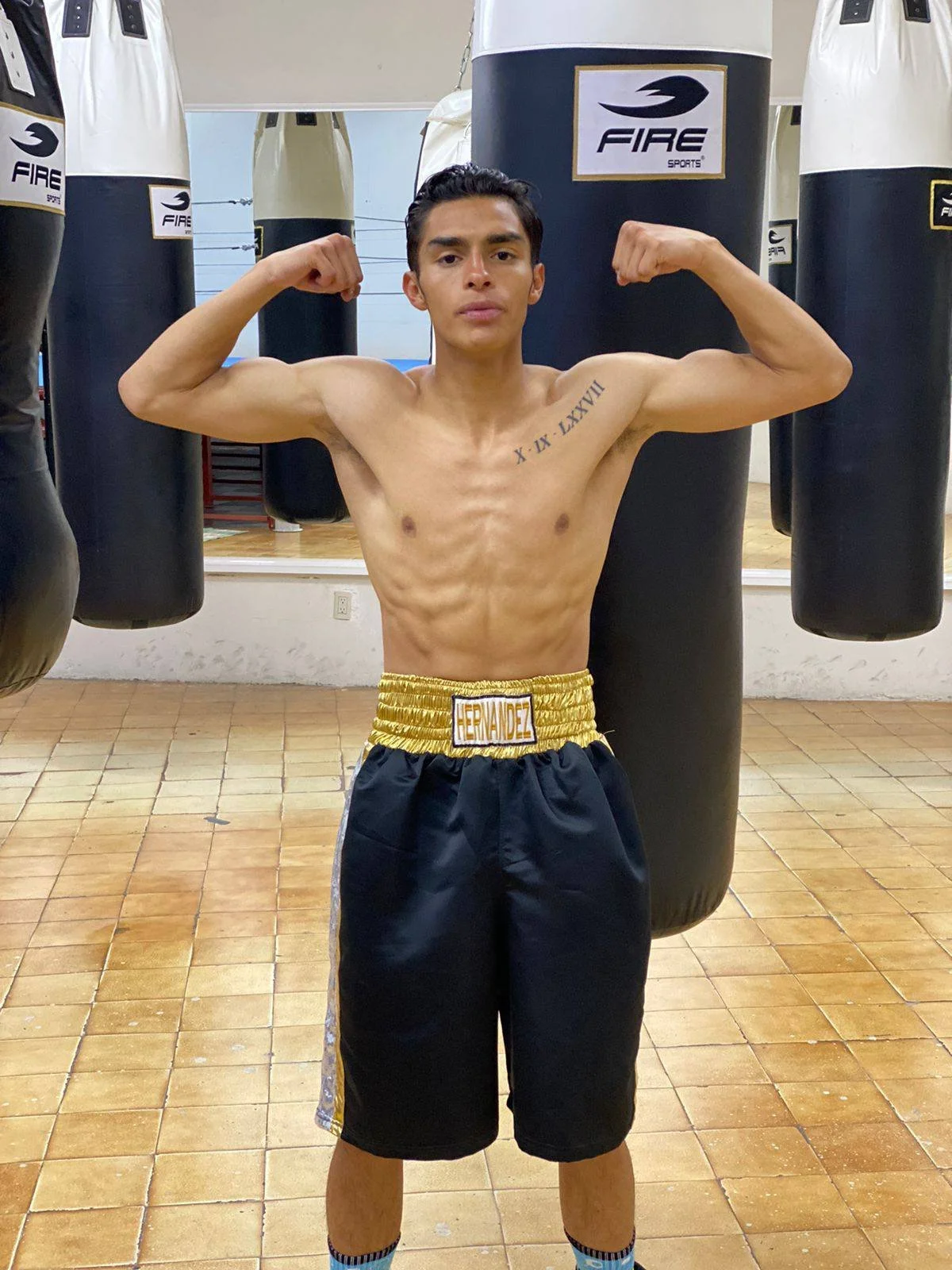 A shirtless young man with tattoos flexing his biceps in boxing shorts in a gym surrounded by punching bags.