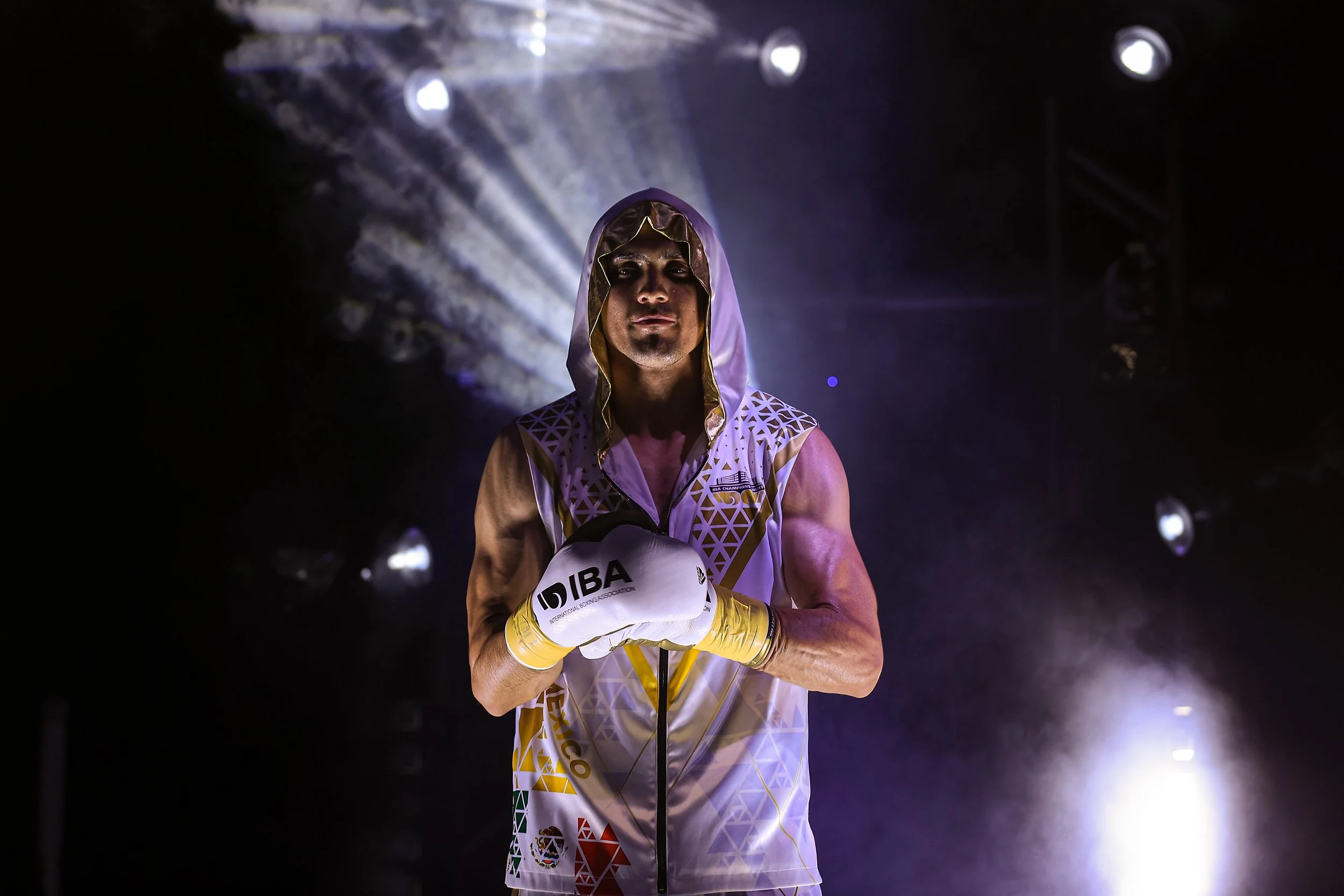 A male boxer in a sleeveless boxing robe with a hood, wearing yellow boxing gloves, standing in a dark arena with spotlight effects behind him, looking confidently at the camera.