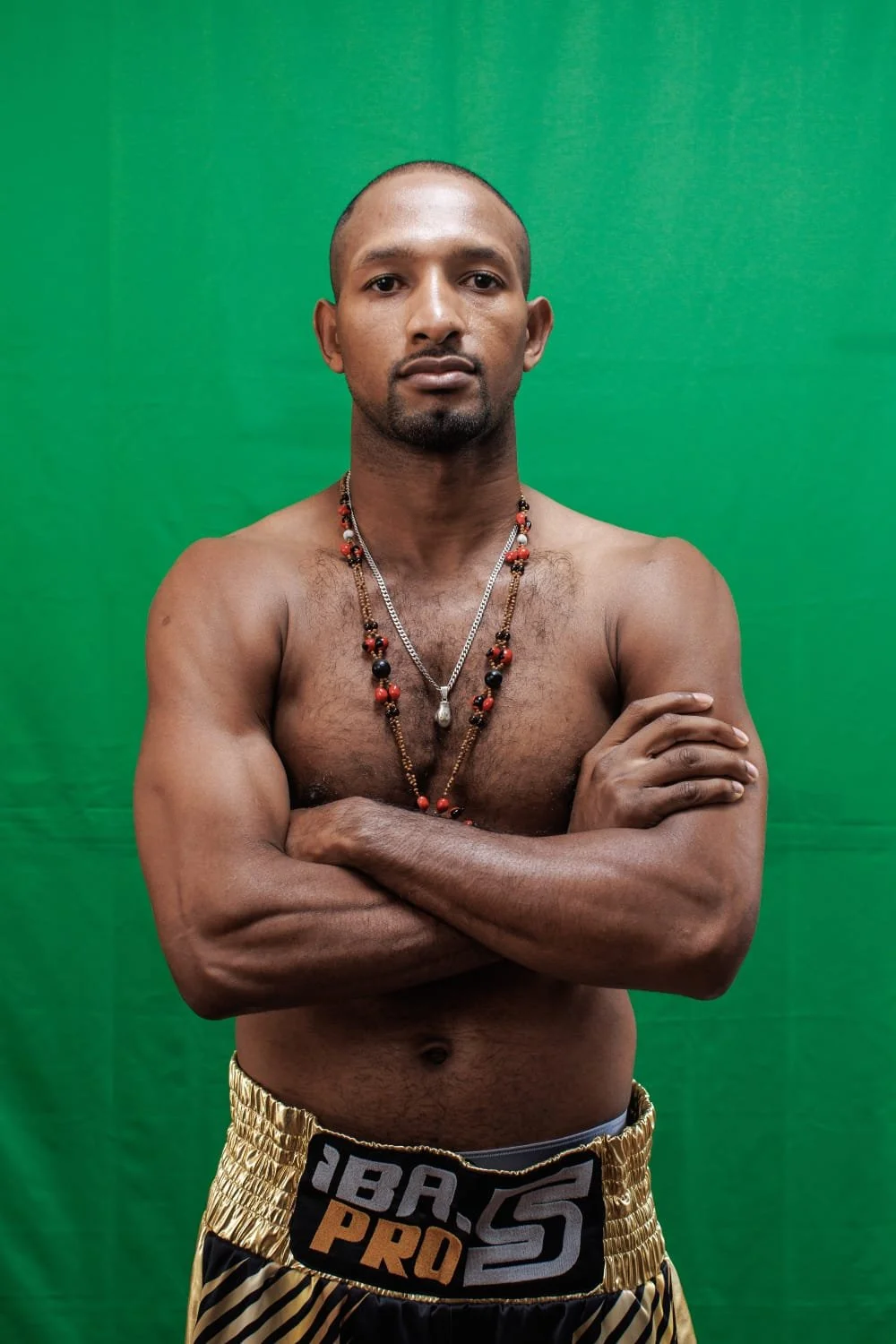 A shirtless man with crossed arms wearing gold and black patterned athletic shorts, multiple beaded necklaces, standing against a green background.