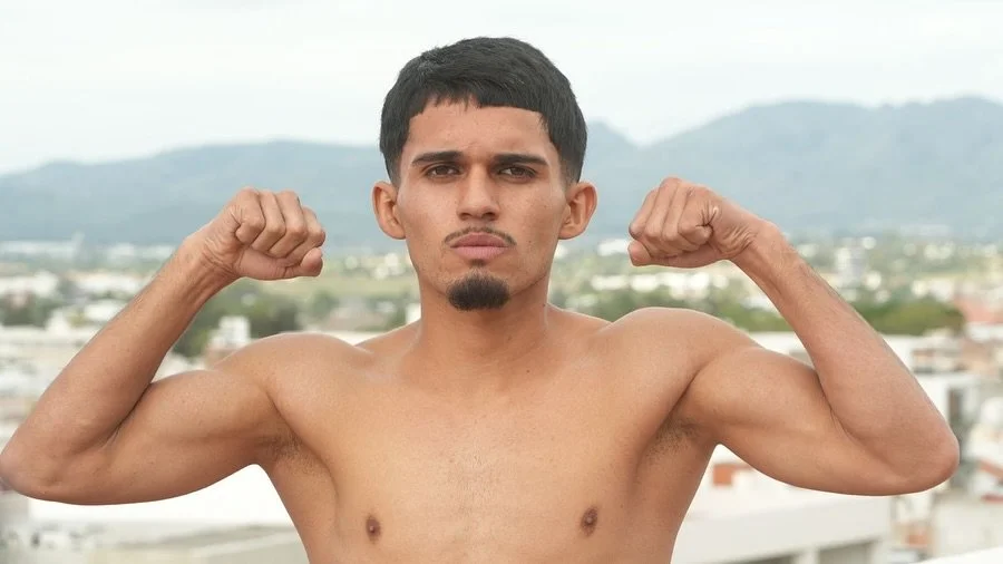 A shirtless young man flexing his biceps outdoors with a cityscape and mountains in the background.