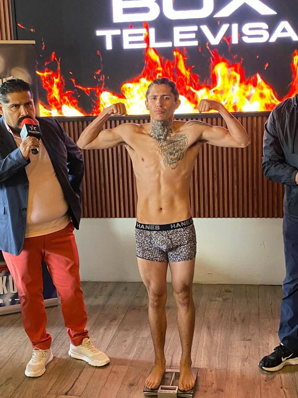 A shirtless man with tattoos on his chest and neck, wearing patterned boxer briefs, stands on a scale, flexing his arms. To his sides are two other men, one holding a microphone. A digital sign behind them displays the words "BOX TELEVISA" with flames in the background.