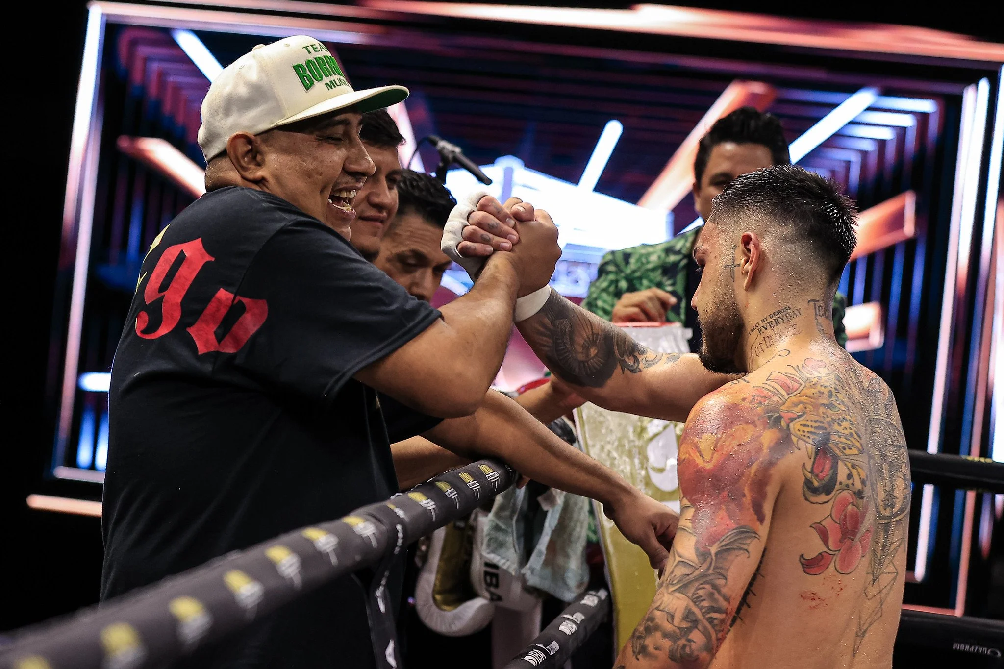 Two men arm wrestling inside a boxing ring, smiling and shaking hands, with other men watching in the background.