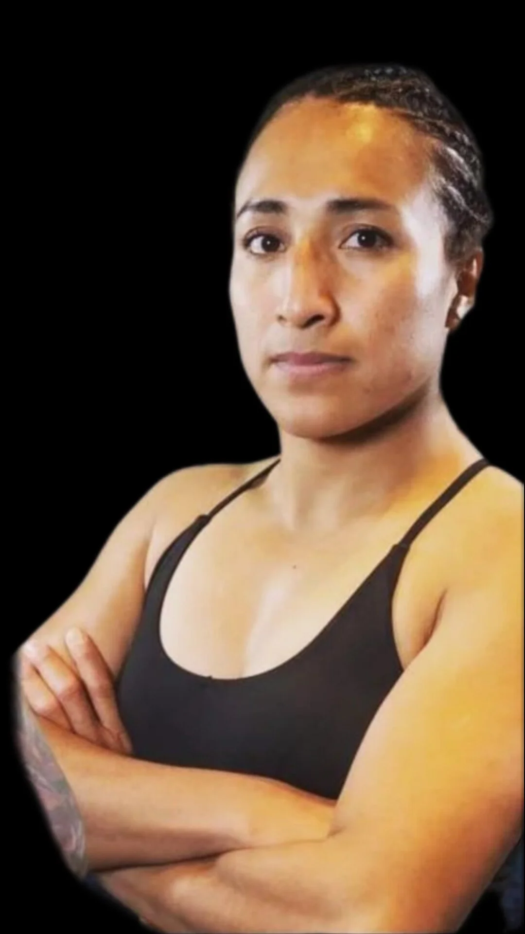 A woman with braided hair wearing a black tank top, standing with arms crossed against a black background.