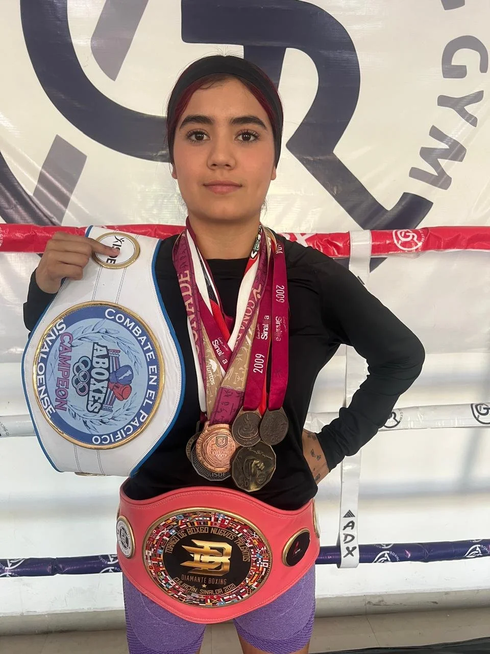 A young female boxer standing in a boxing gym with medals around her neck, holding a large championship belt, wearing a pink championship belt around her waist. She has a confident expression, wearing a black shirt, purple leggings, and a black headband. Medals and a logo are visible in the background.