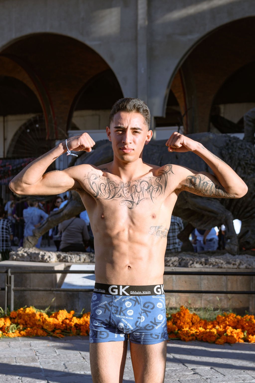 A shirtless young man with tattoos flexing his biceps outdoors, with arches and people in the background, and orange flowers on the ground.