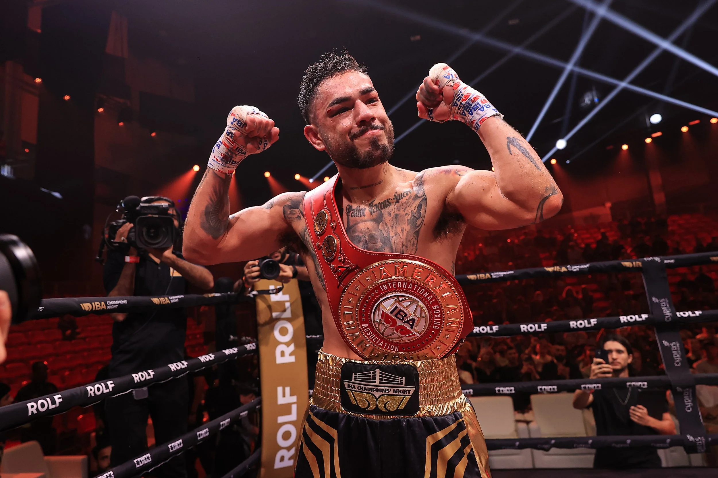 A male boxer inside a boxing ring, flexing his biceps, wearing a red championship belt over his shoulder and gold shorts, surrounded by photographers and spectators.