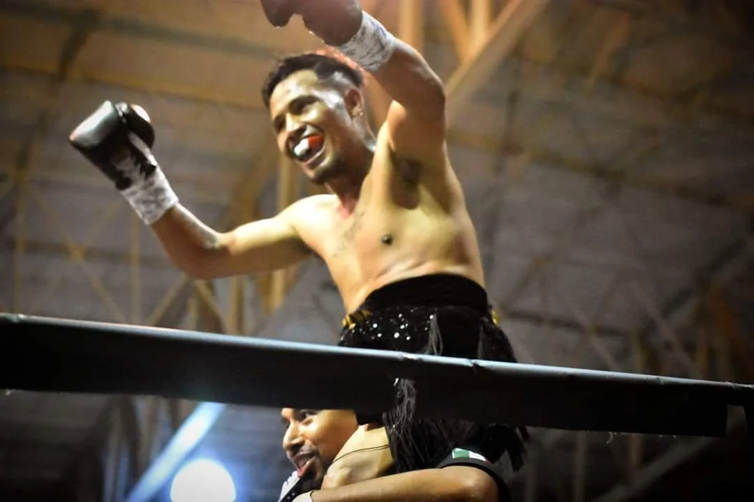 A shirtless male MMA fighter celebrating in the ring, with his arms raised and a smile, while holding boxing gloves in both hands.