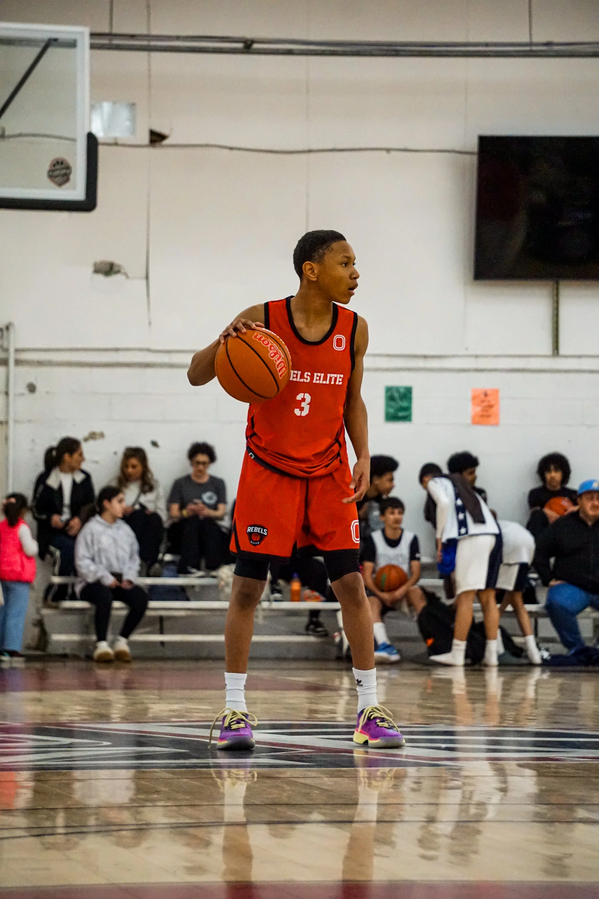 A young basketball player dribbling the ball on an indoor court during a game, with spectators and officials watching.