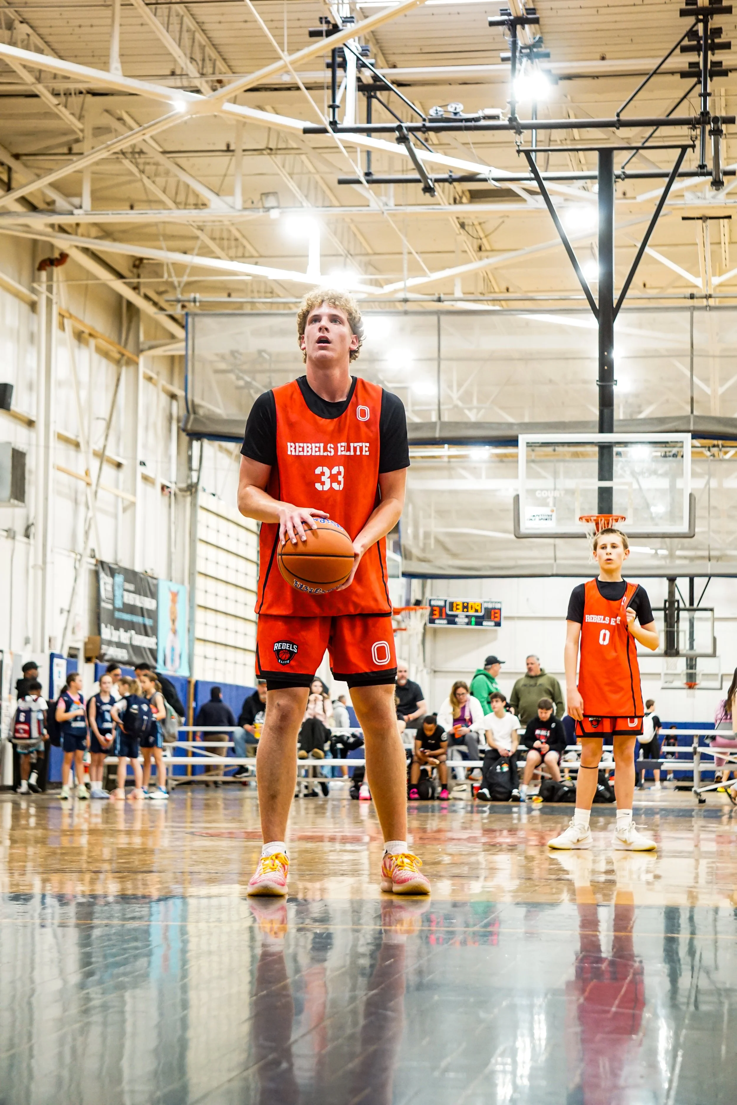 A young male basketball player walking on an indoor court, wearing a white and black jersey with red accents, number 3, and black shorts with red and white details.