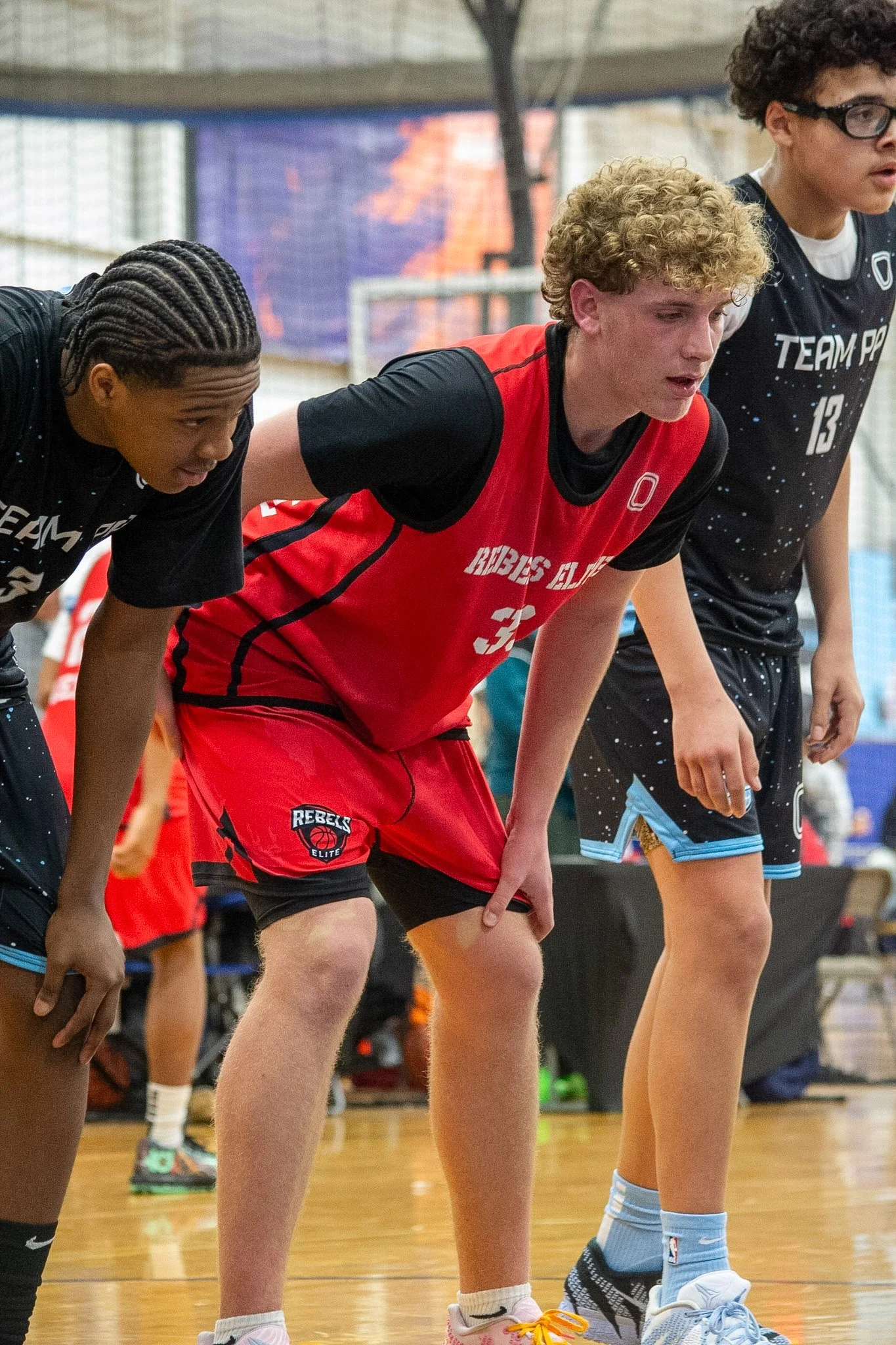 A basketball player wearing a black jersey with red and white accents, number 18, is jumping towards the basketball hoop to make a shot. Other players and spectators are visible in the background inside a gymnasium.