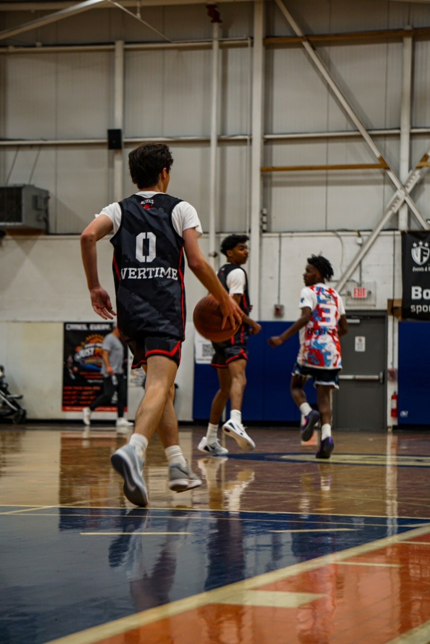 A basketball player in a red shirt and beige shorts with red and black details jumping to make a slam dunk at an indoor basketball court.